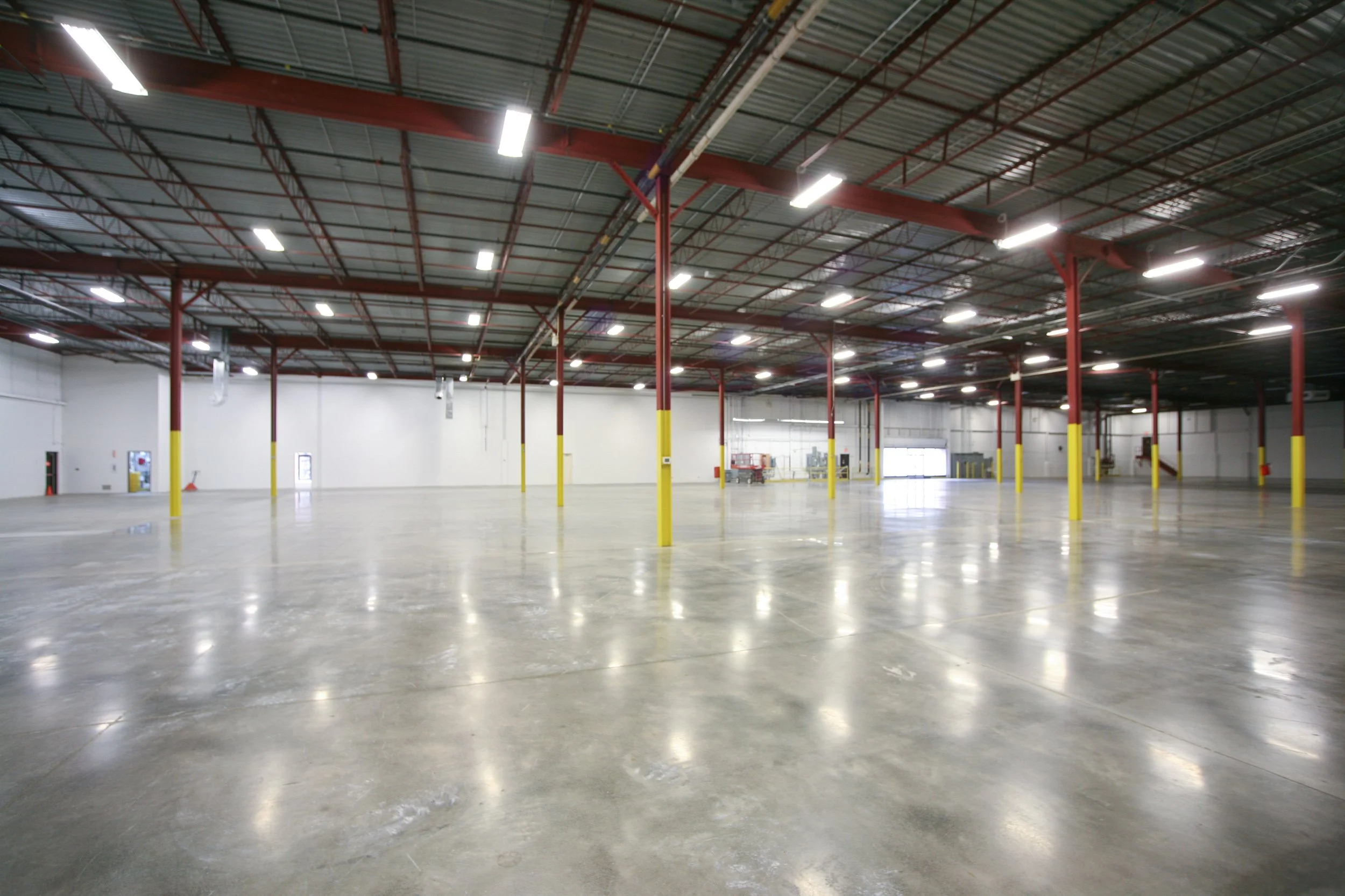 Empty warehouse with a polished concrete floor, red support beams, and bright overhead lighting.