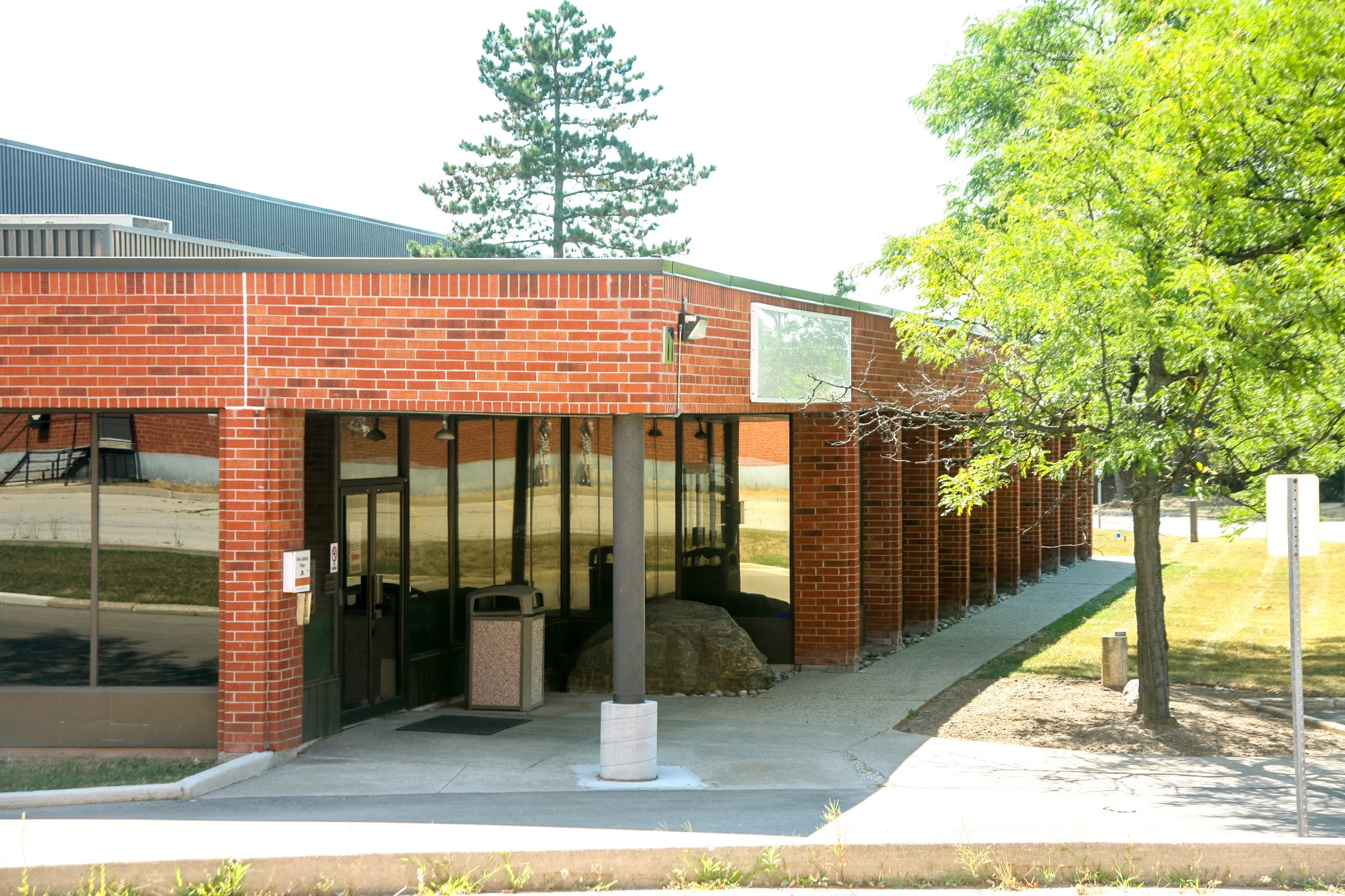 A brick building with multiple glass doors and windows, a sidewalk, trees, and a blue sky.