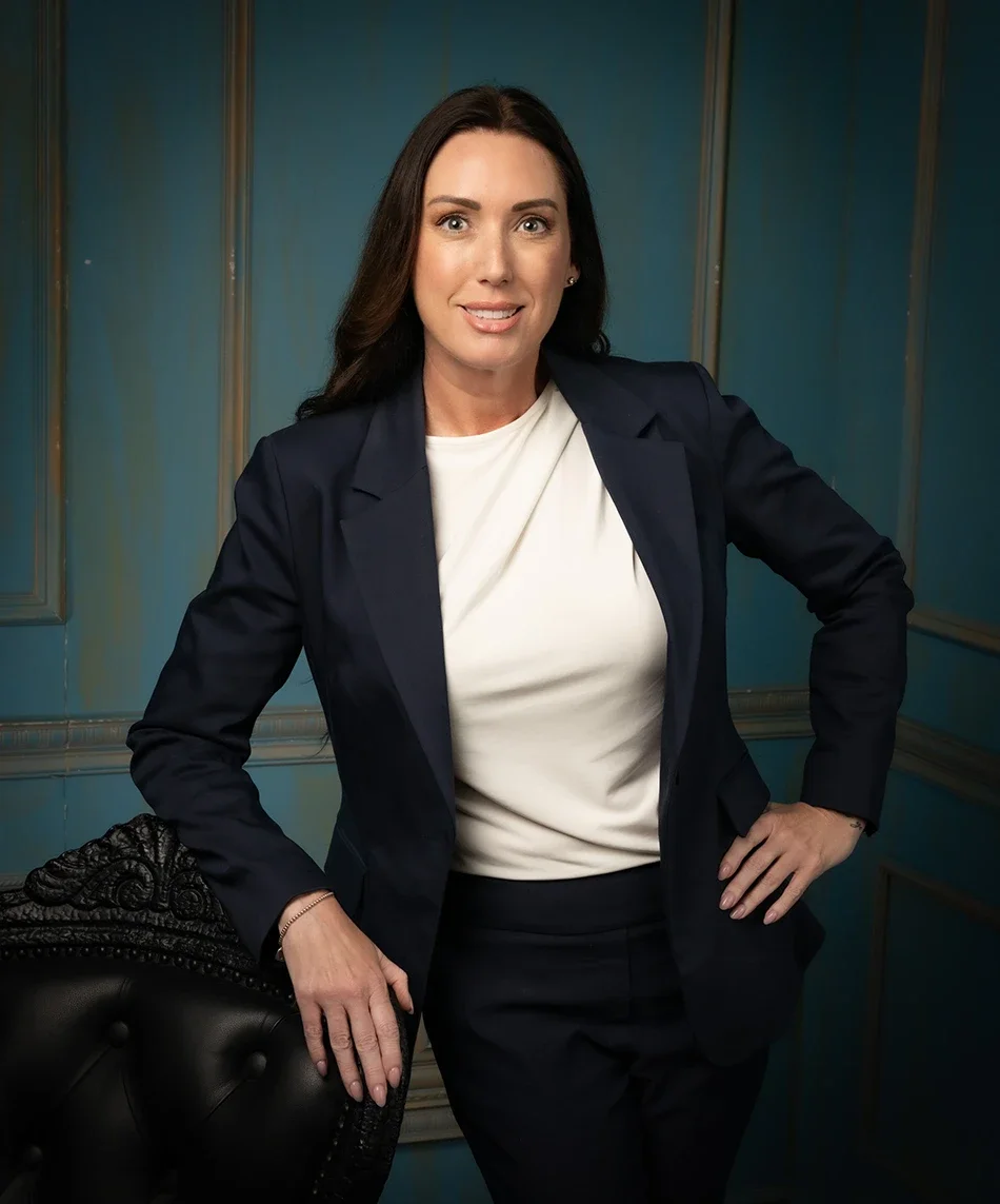 Woman with long brown hair in black outfit standing next to a black chair in a dimly lit room with gray paneled walls.