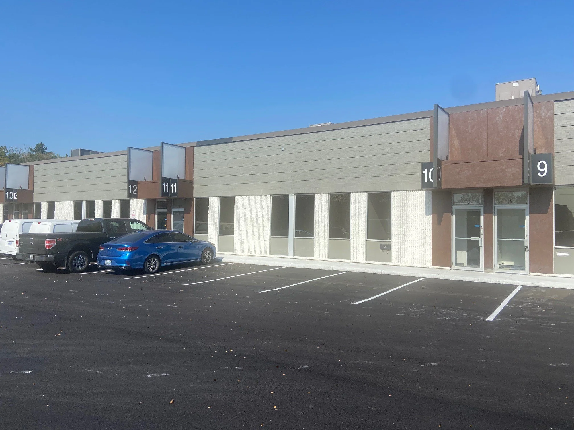 View of a commercial building with numbered sections and a parking lot filled with several cars. Clear blue sky overhead.