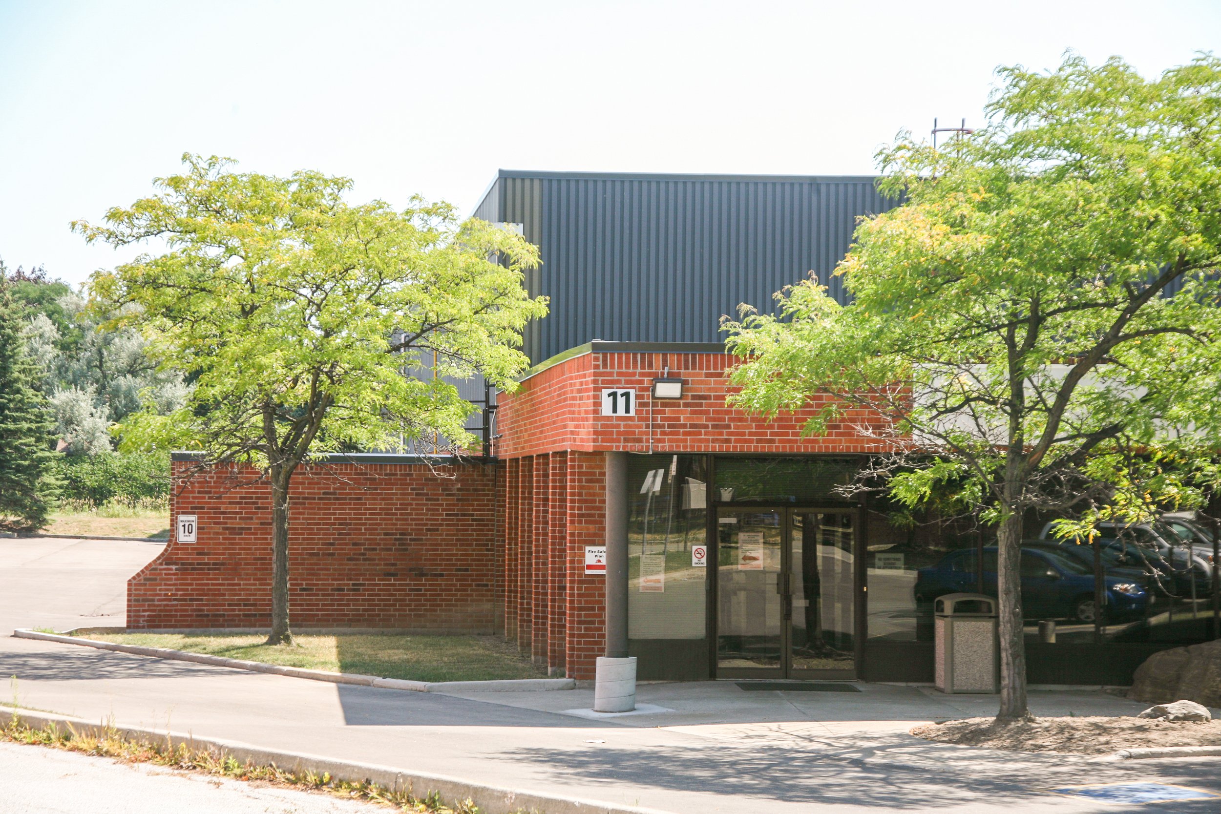 A brick building with the number 11 on it, surrounded by green trees, with a bus stop in front, and parking lot behind, under a clear sky.