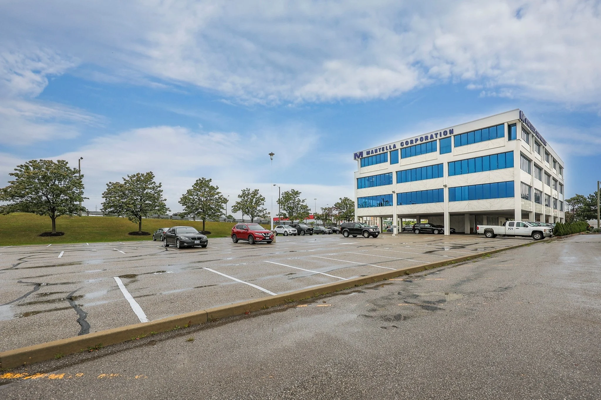 A parking lot with a few parked cars, a grassy area with trees, and a large white office building with blue windows labeled "Mantella Corporation" in the background under a partly cloudy sky.