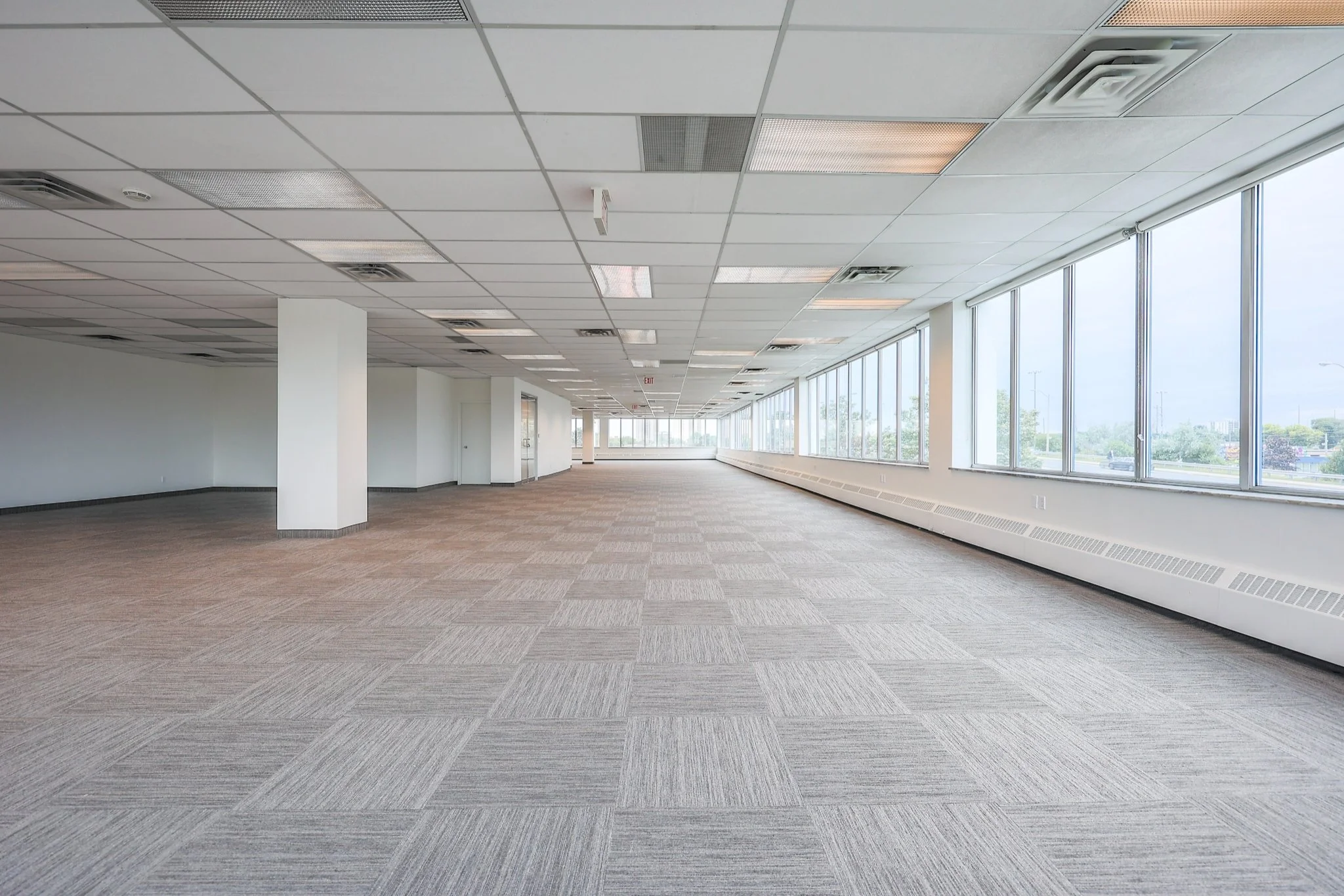 Empty office space with large windows, white walls, gray carpet, white ceiling with rectangular light panels, and visible air vents.