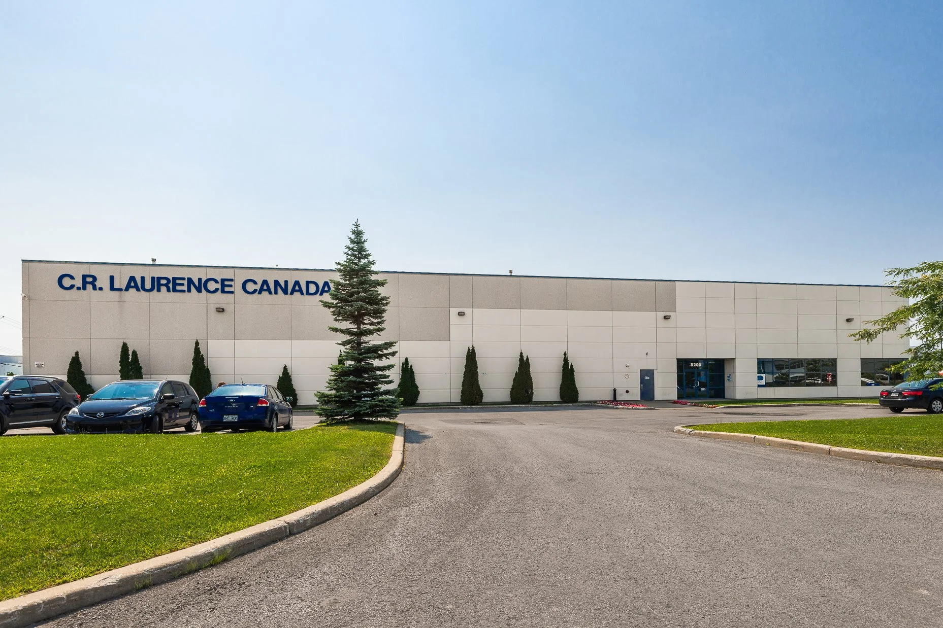 Commercial building with the sign 'C.R. LAURENCE CANADA', parking lot with several cars and green trees in front, clear blue sky.