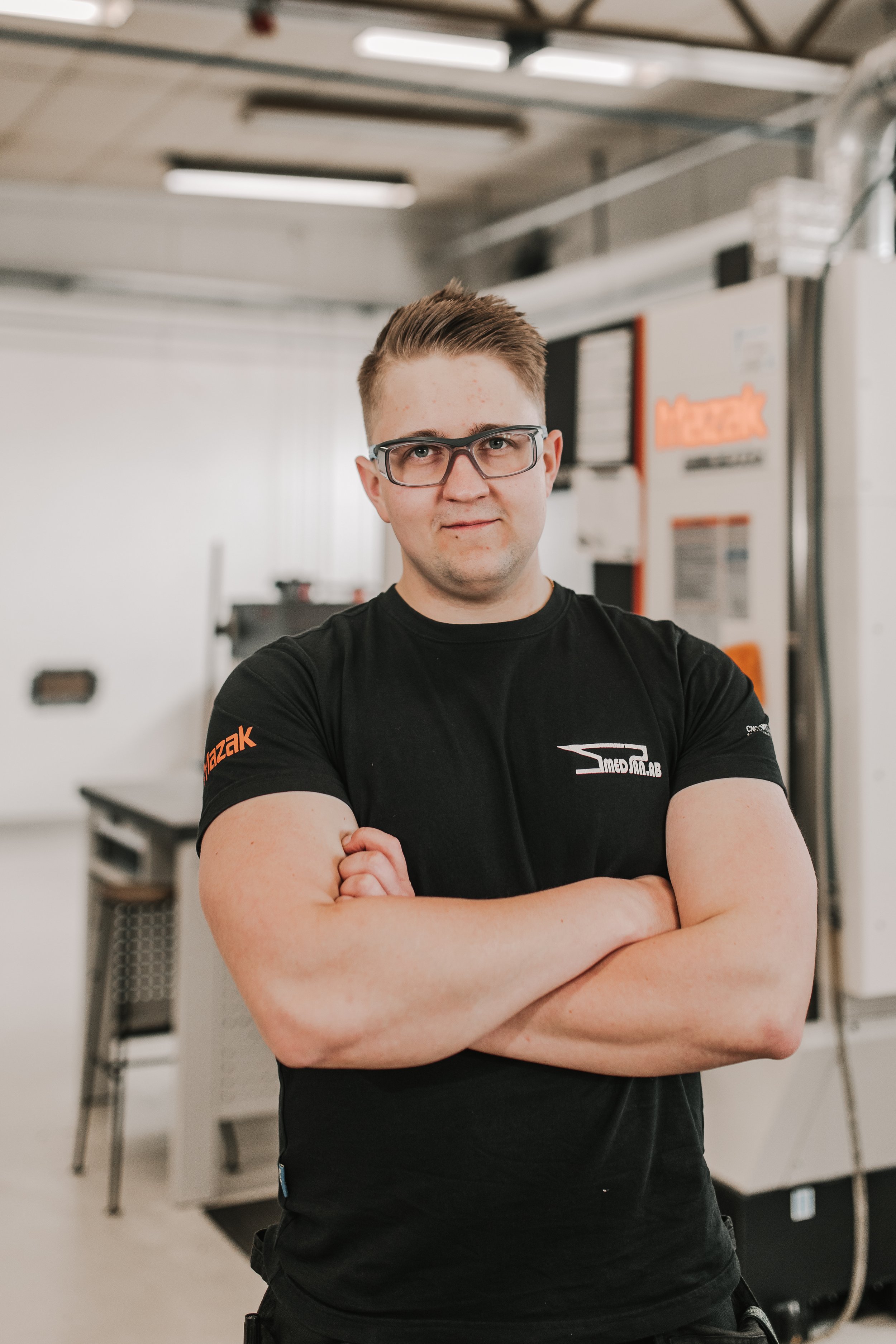 En ung man står med armar korsade i en industrimiljö, bär svart t-shirt med logotyper, glasögon och har kort, uppklippt hår.