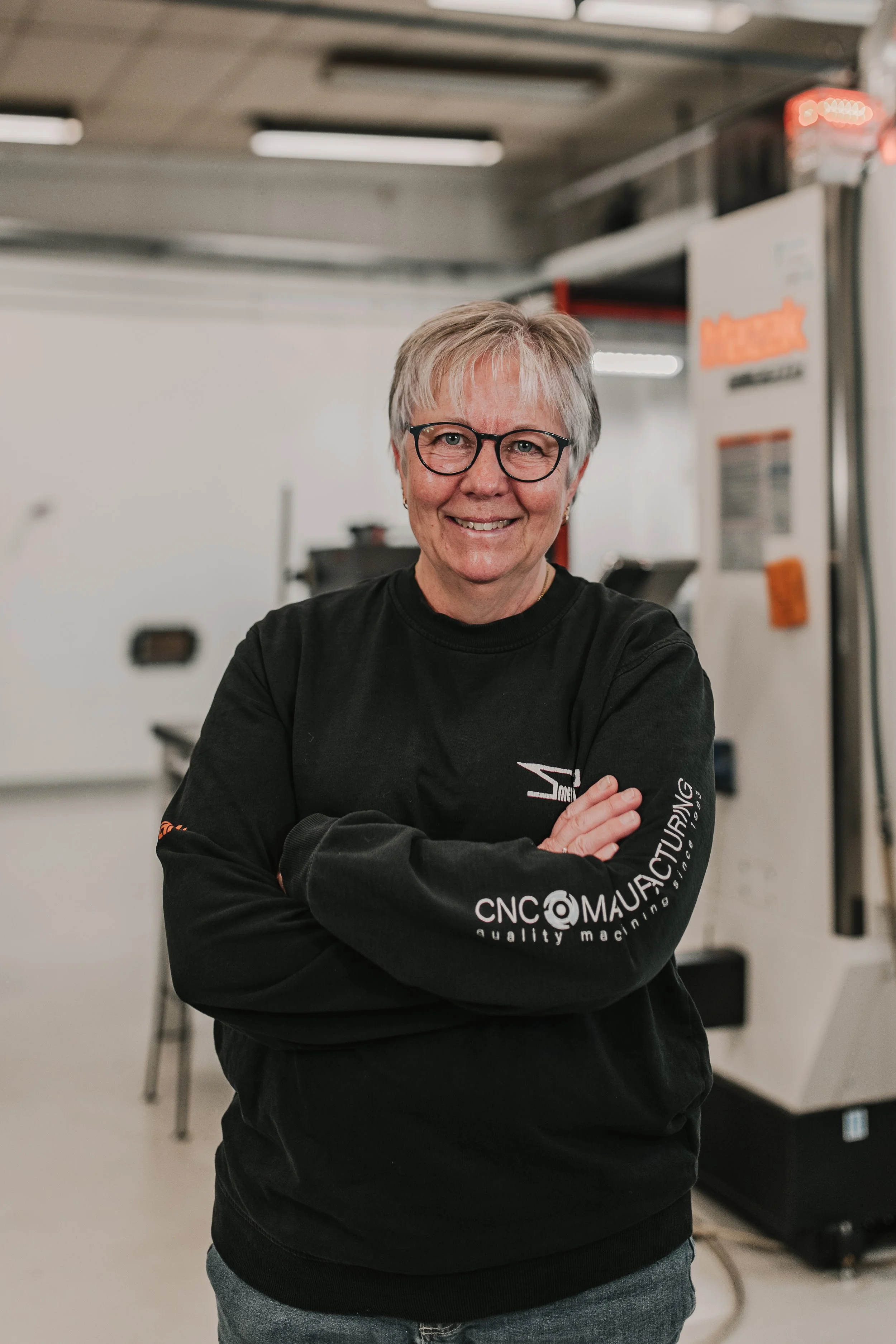 En glad kvinna med grått hår och glasögon står i en industriell miljö, korsar armarna, bär en svart tröja med företagslogotyp.