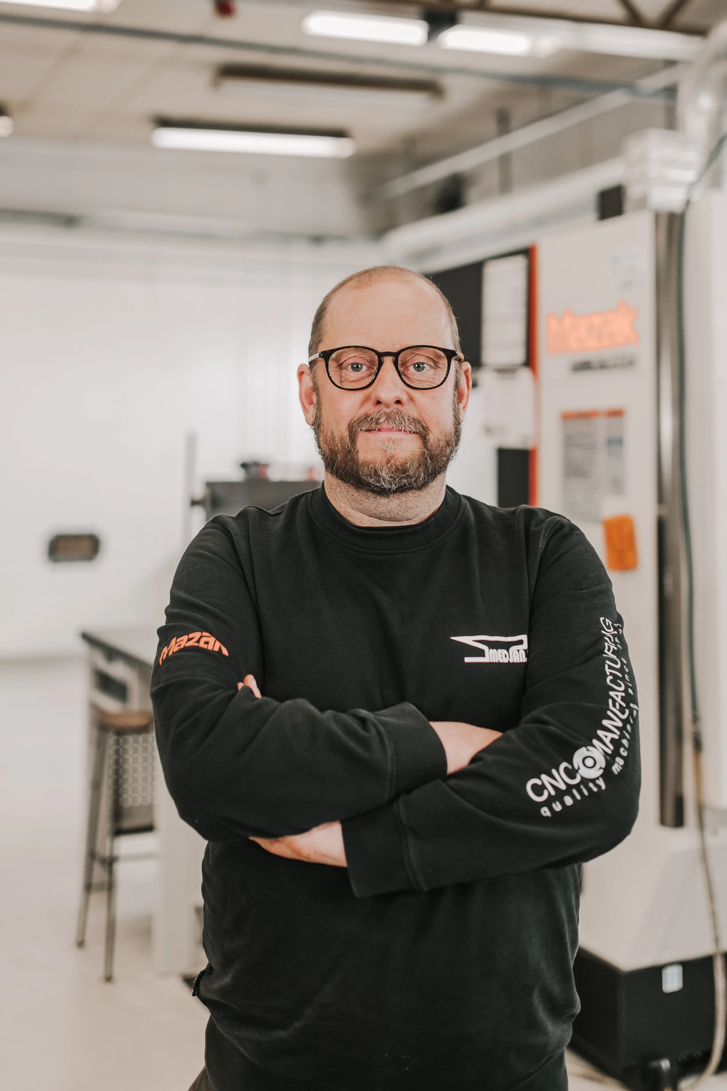 En man med glasögon och skägg, står med armarna i kors i ett industriellt eller verkstadsmiljö, iklädd svart sweatshirt med företagslogotyper.
