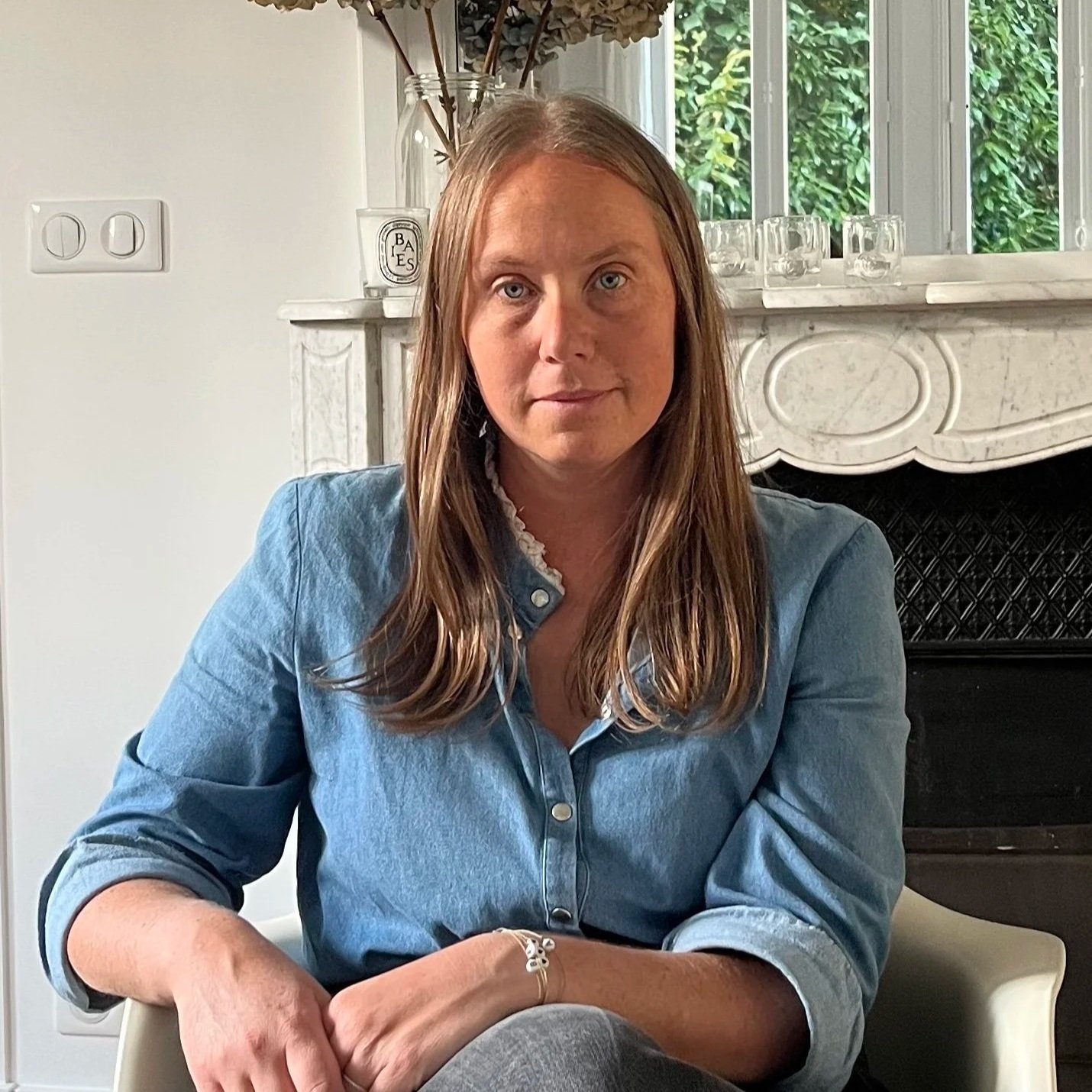 Lucie Mougne, belle femme aux cheveux blonds, portant une chemise en jean bleue, assise dans une pièce avec cheminée en marbre blanche, et une fenêtre derrière avec vue sur des plantes verdoyantes.