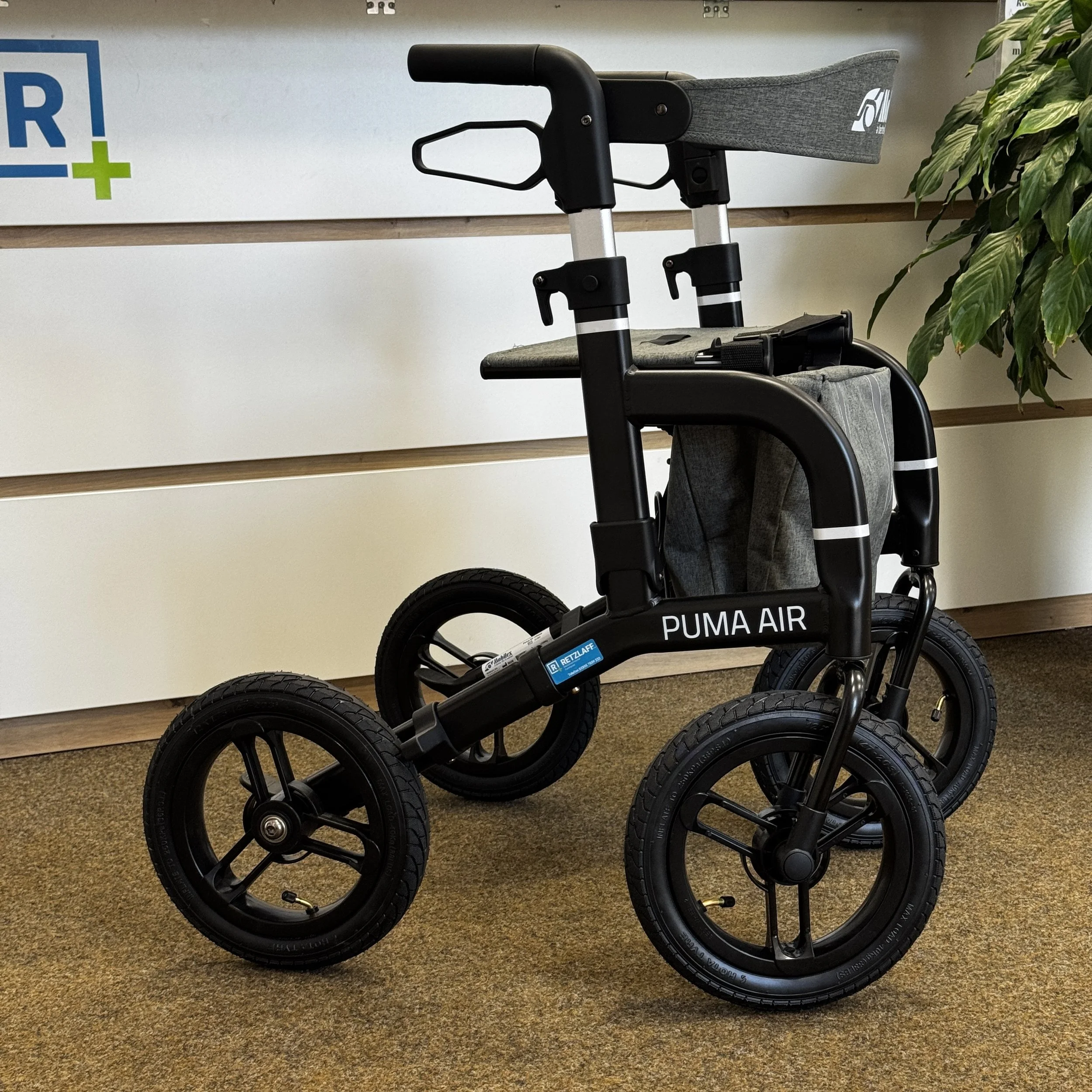 Ein schwarzer Rollstuhl mit den Worten "PUMA AIR" auf dem Rahmen, steht auf einem Teppichboden vor einer weißen Wand mit Holzpanelen.