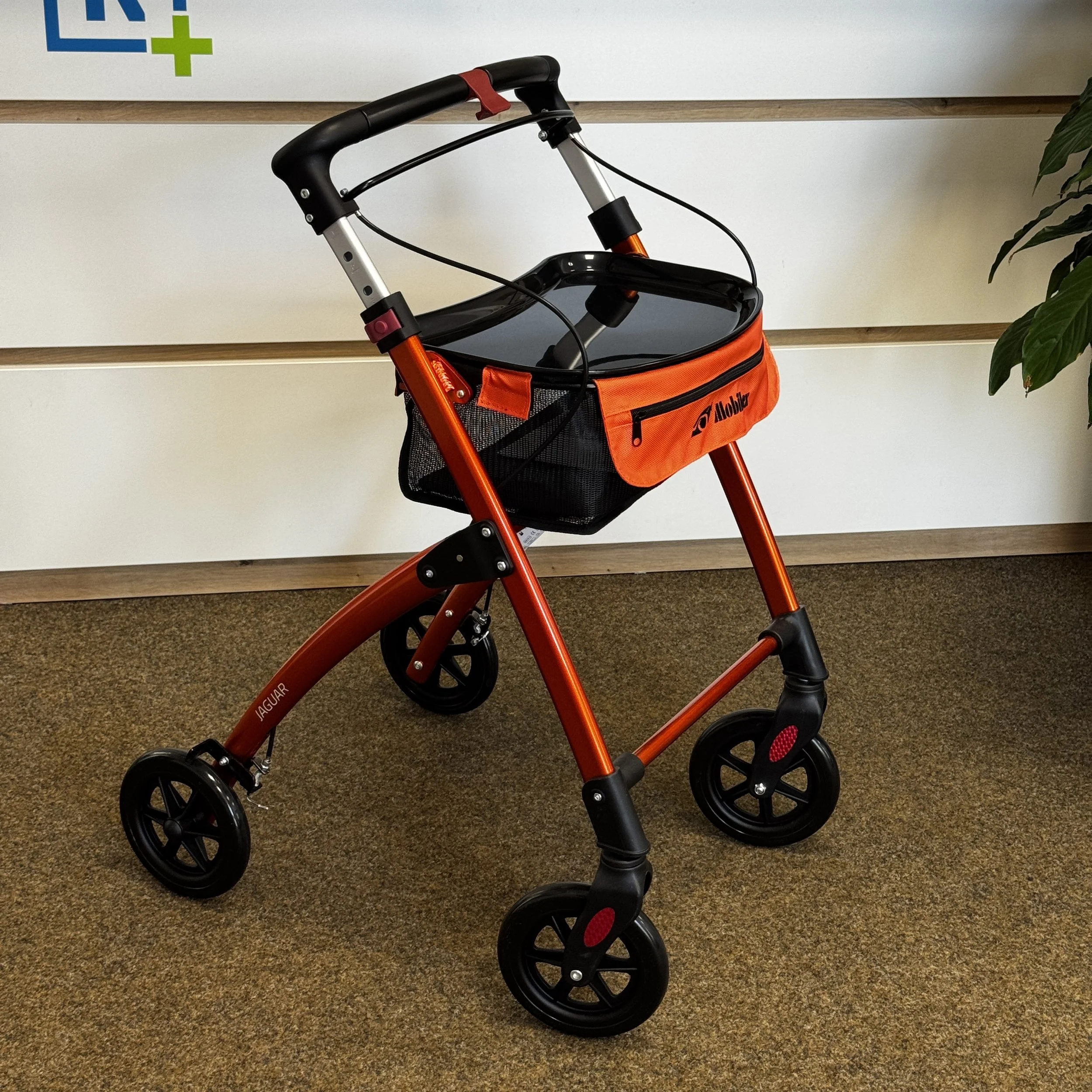 Ein rotes Gehwagen mit schwarzem Korb und orangefarbener Tasche auf glattem Boden vor einer weißbraunen Wand.