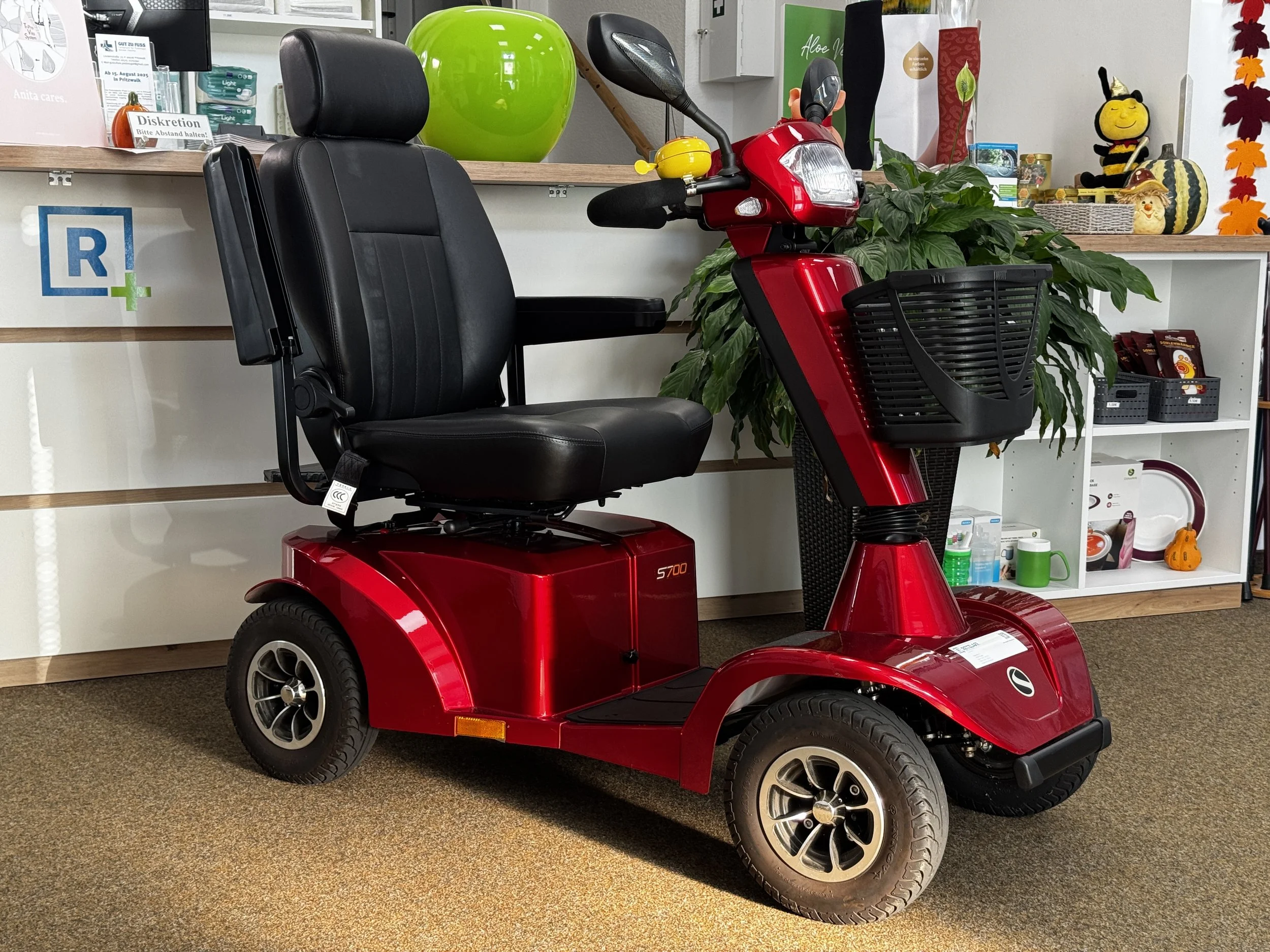 Ein roter elektrischer Dreiradrollstuhl mit schwarzem Sitz und Lenkrad, in einem Innenraum mit Dekorationen und einem Regal im Hintergrund.