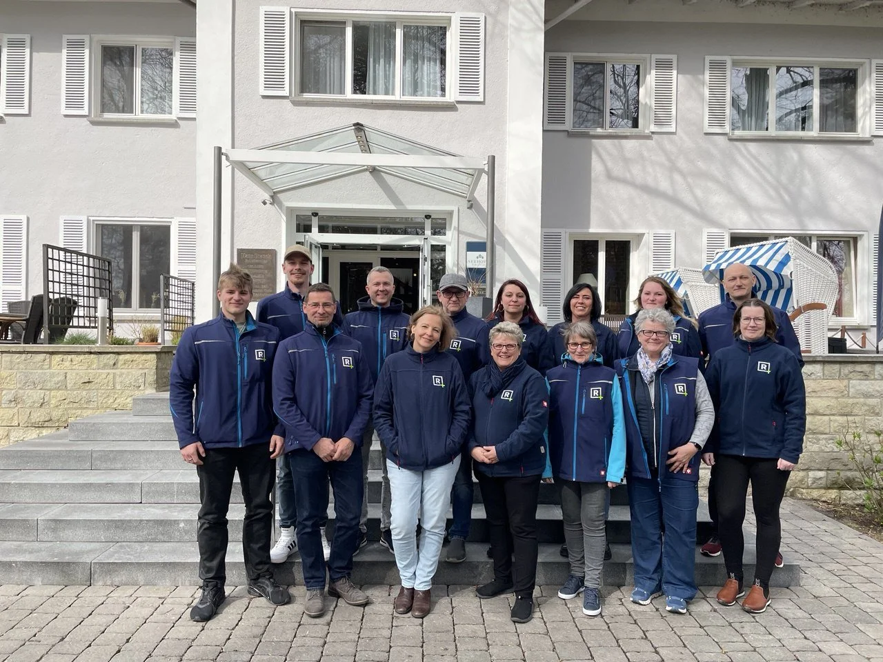 Gruppe von Menschen in blauen Jacken steht vor einem weißen Gebäude mit mehreren Fenstern.