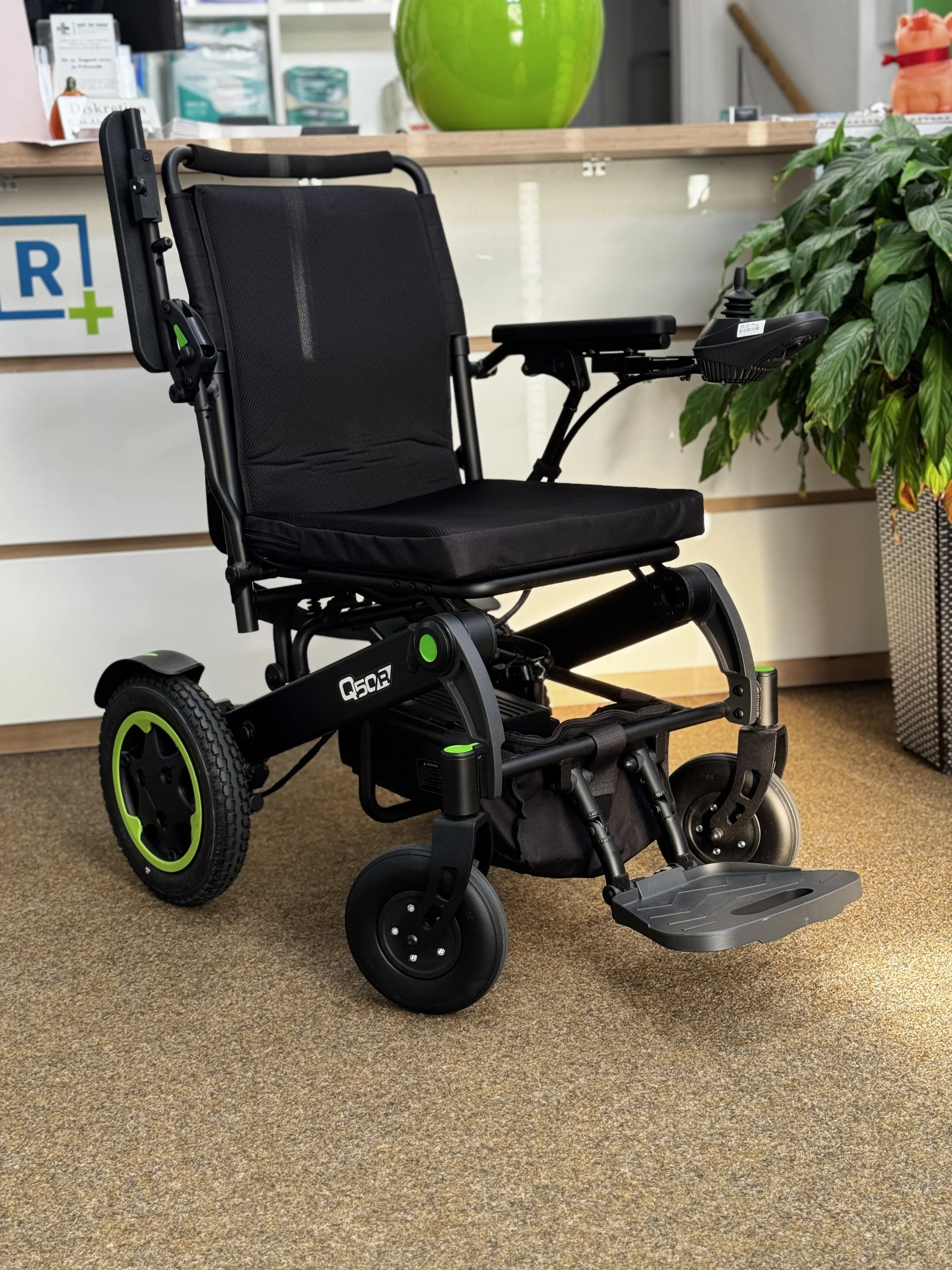 Elektrischer Rollstuhl auf Teppichboden in einem Wartebereich, mit einem Empfangstresen im Hintergrund und einer grünen Pflanze auf der rechten Seite.