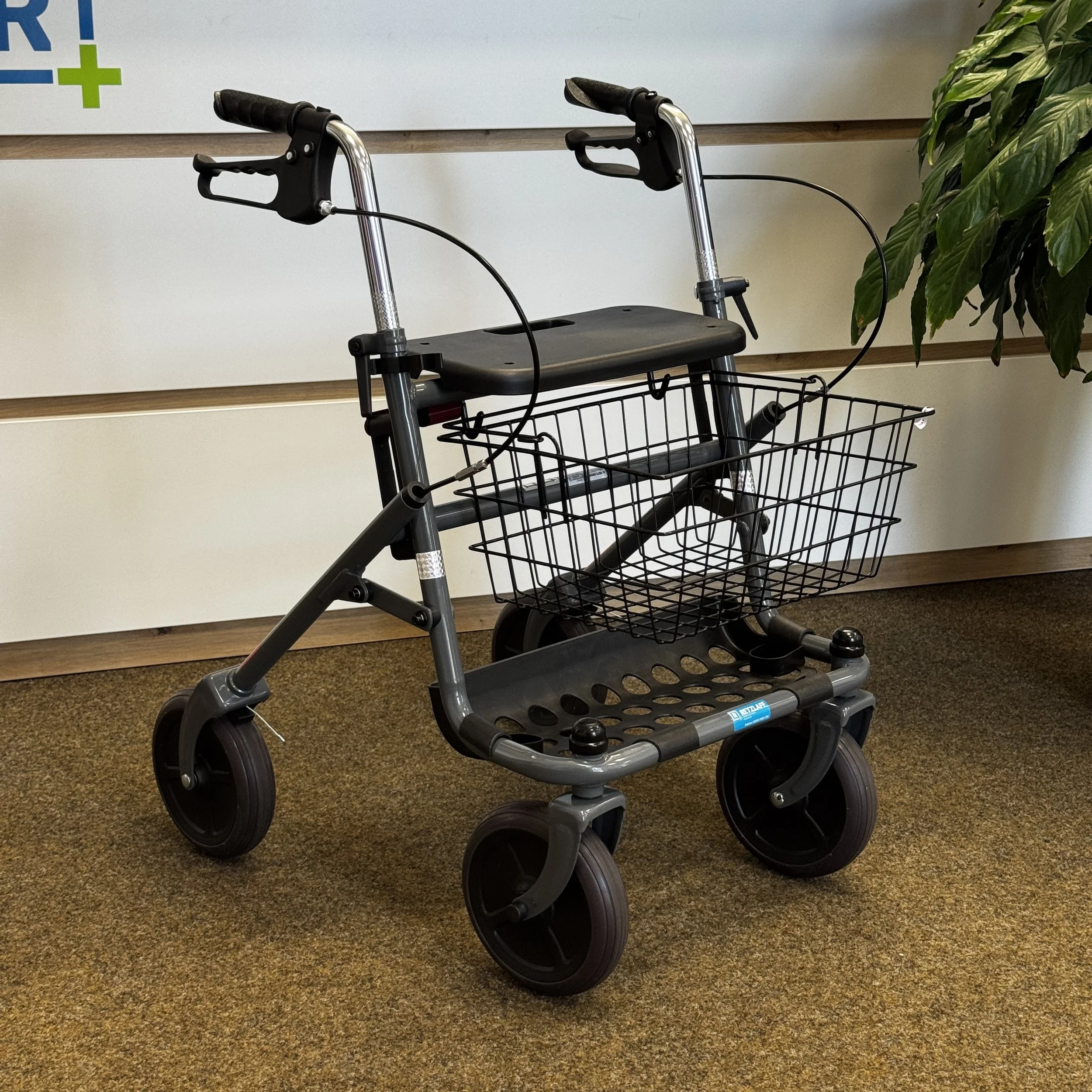 Ein rotes und schwarzes Rollator mit einer großen Einkaufskorbtasche vor einem weißen Schrank und einer Pflanze