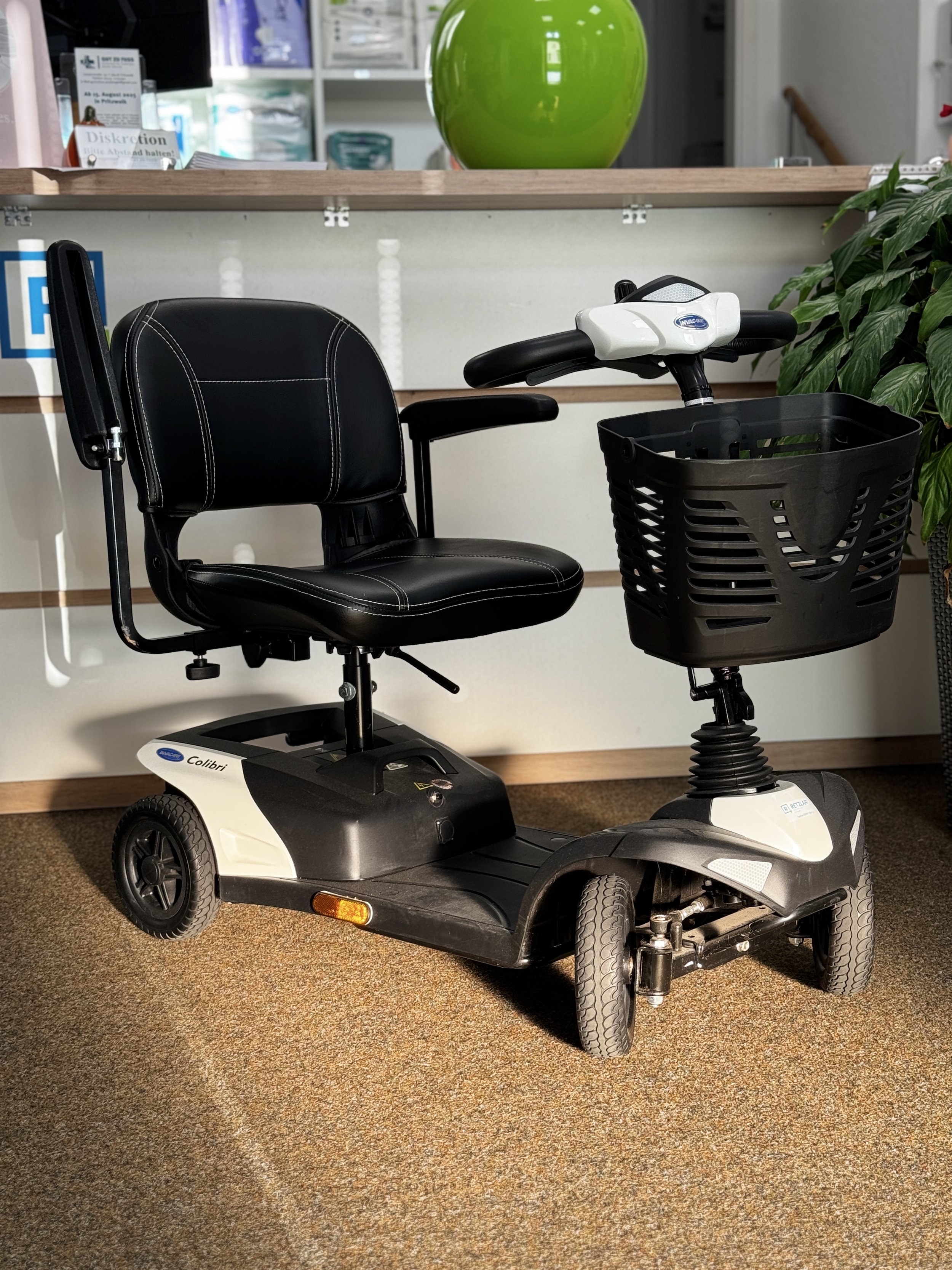 Elektrischer Rollstuhl mit Sitz, Armlehnen und Einkaufskorb, auf braunem Teppichboden, im Hintergrund grüne Pflanze und weiß-gestreifte Wand.