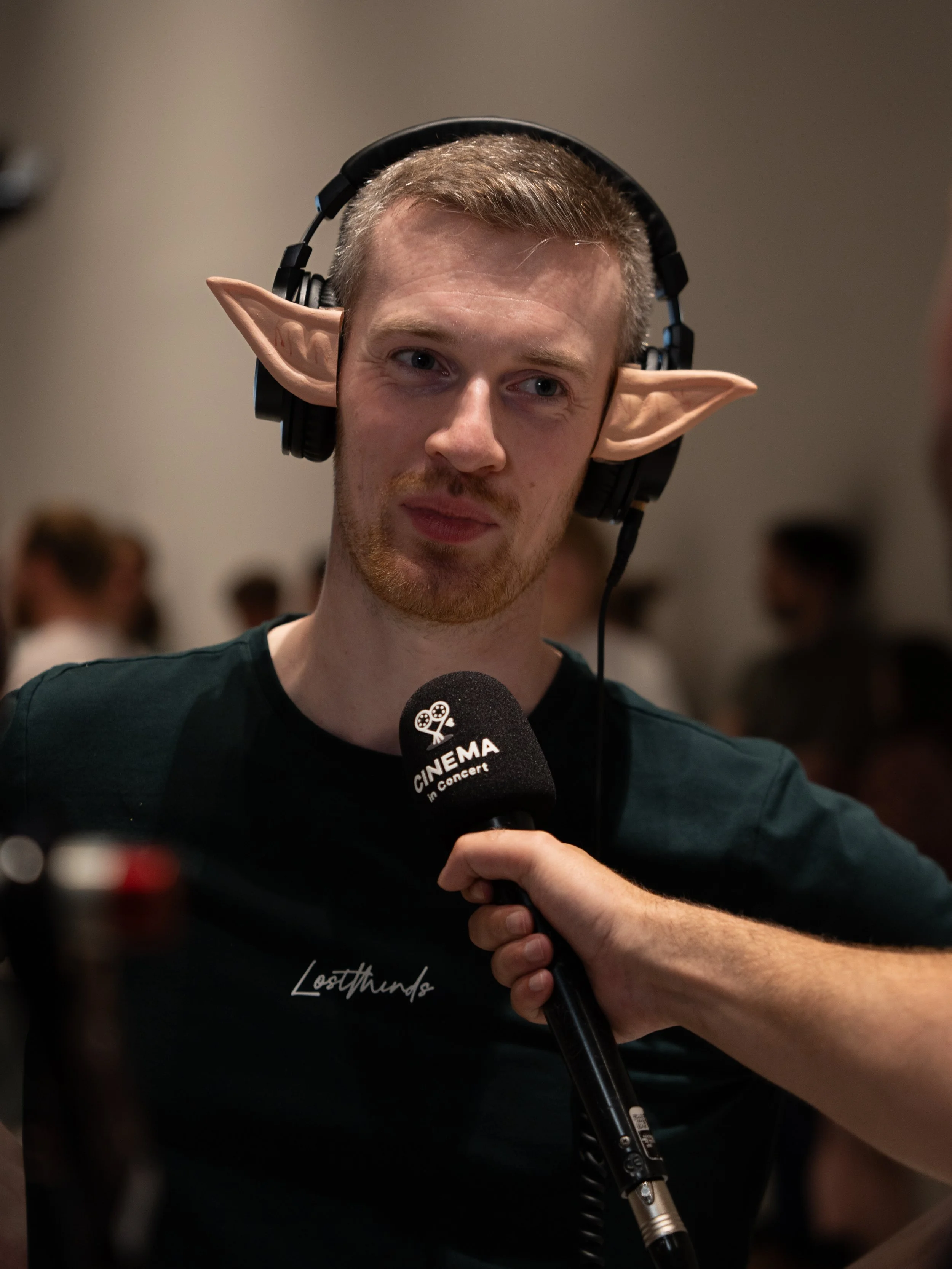 A person wearing large headphones with novelty elf ears attached, speaking into a microphone labeled 'Cinema in Concert.' The person has a neutral expression and is in an indoor setting with blurred people in the background.