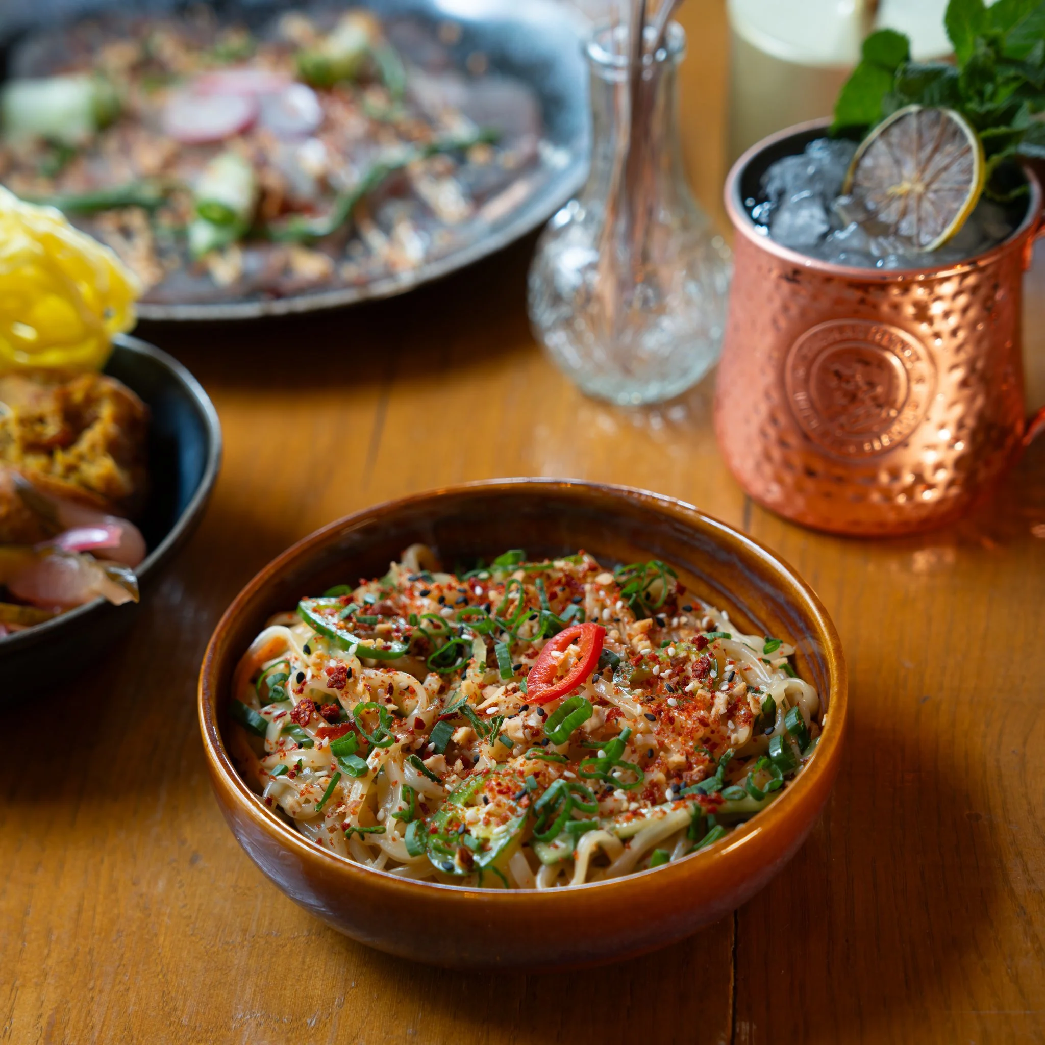 A bowl of noodles garnished with herbs, chili slices, and spices on a wooden table, accompanied by a copper mug with ice and a slice of lemon, and other plates of food blurred in the background.