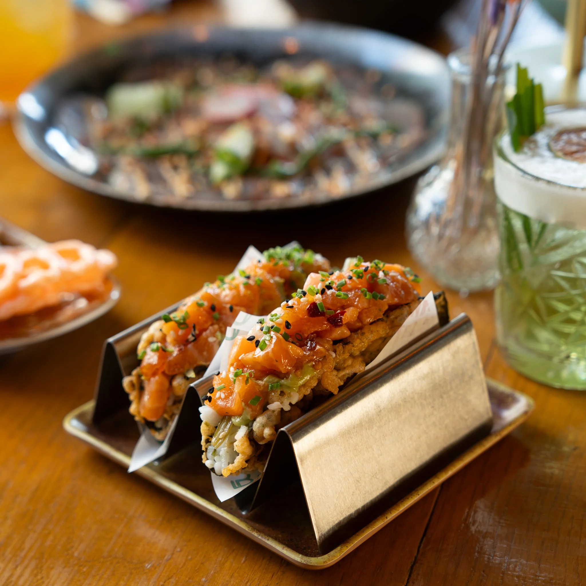 Sushi tacos with salmon, avocado, and garnish in a metal holder, surrounded by other dishes and a cocktail on a wooden table.