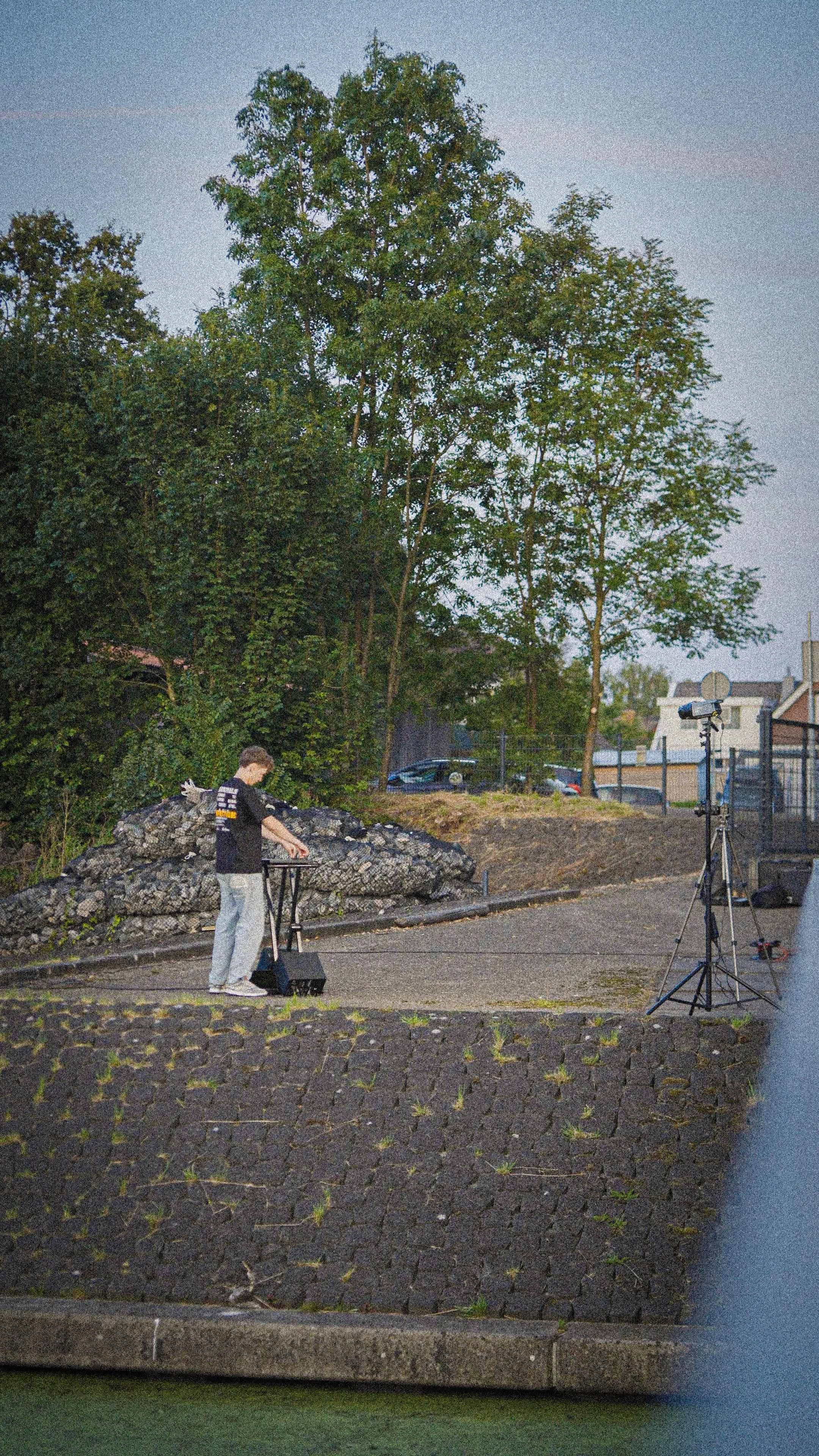 Person setting up equipment outdoors near a paved path and green trees, with a lighting stand nearby.