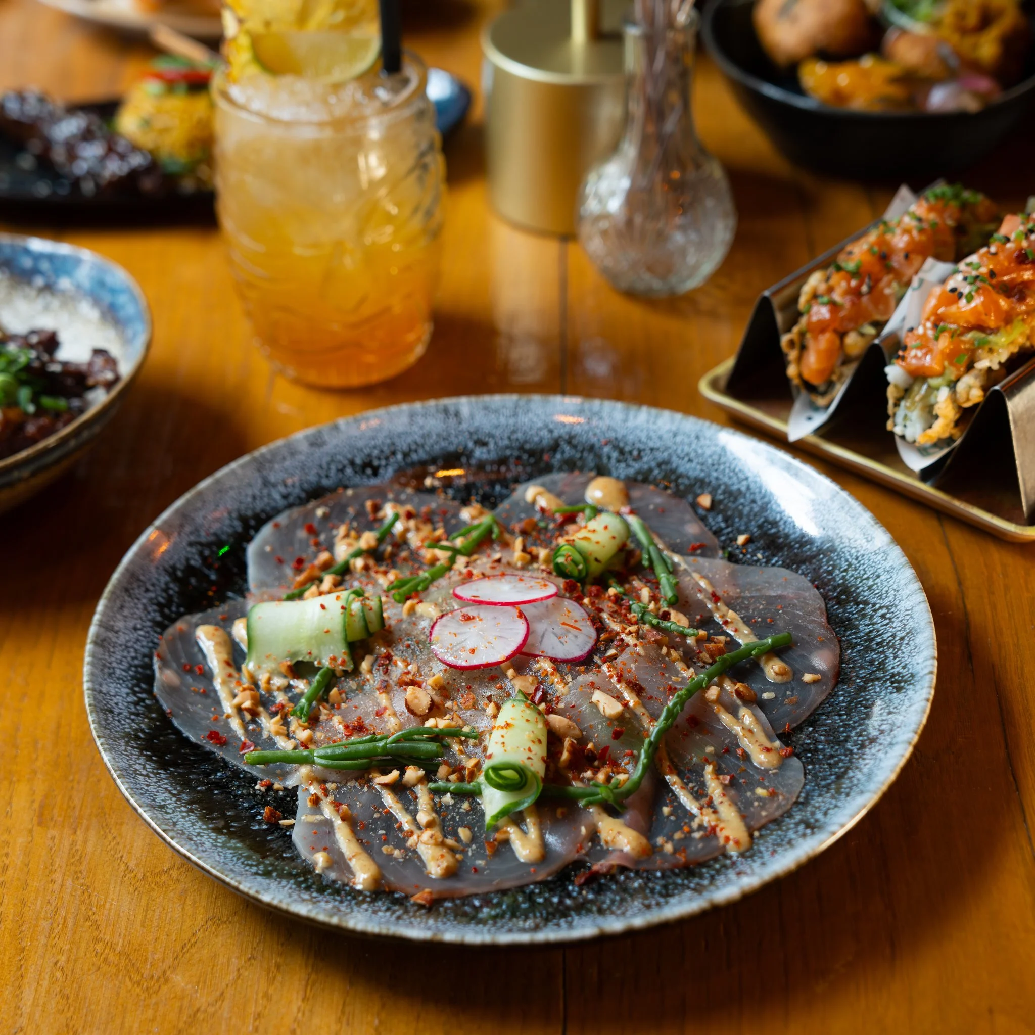 Close-up of a gourmet dish featuring thinly sliced meat garnished with radishes, cucumber, and green beans on a dark plate, accompanied by a tropical cocktail and tacos in the background.