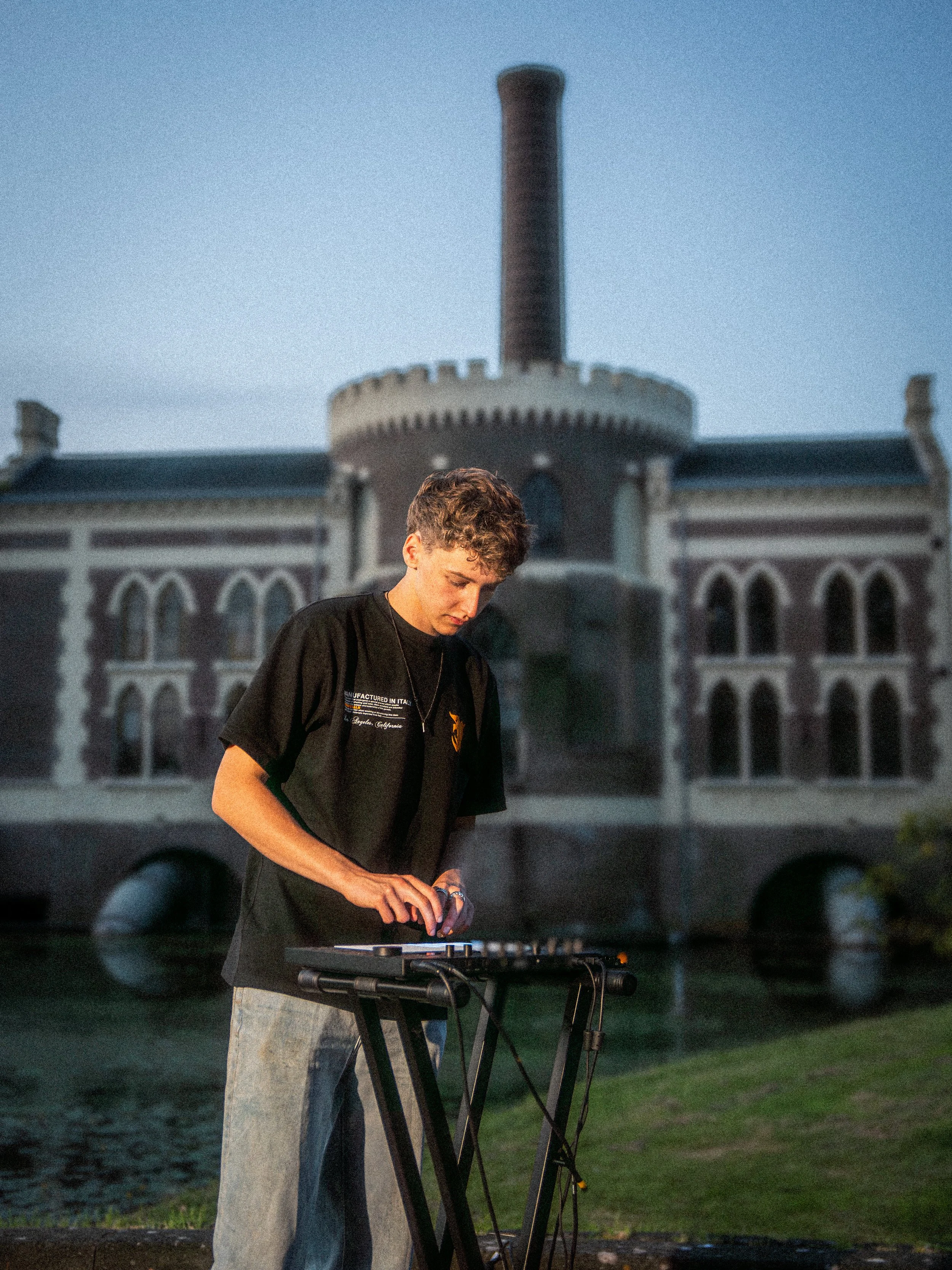 A young man wearing a black t-shirt and jeans is playing music on a keyboard in front of a historic building with a tall chimney. The building has arched windows and a water feature in front.