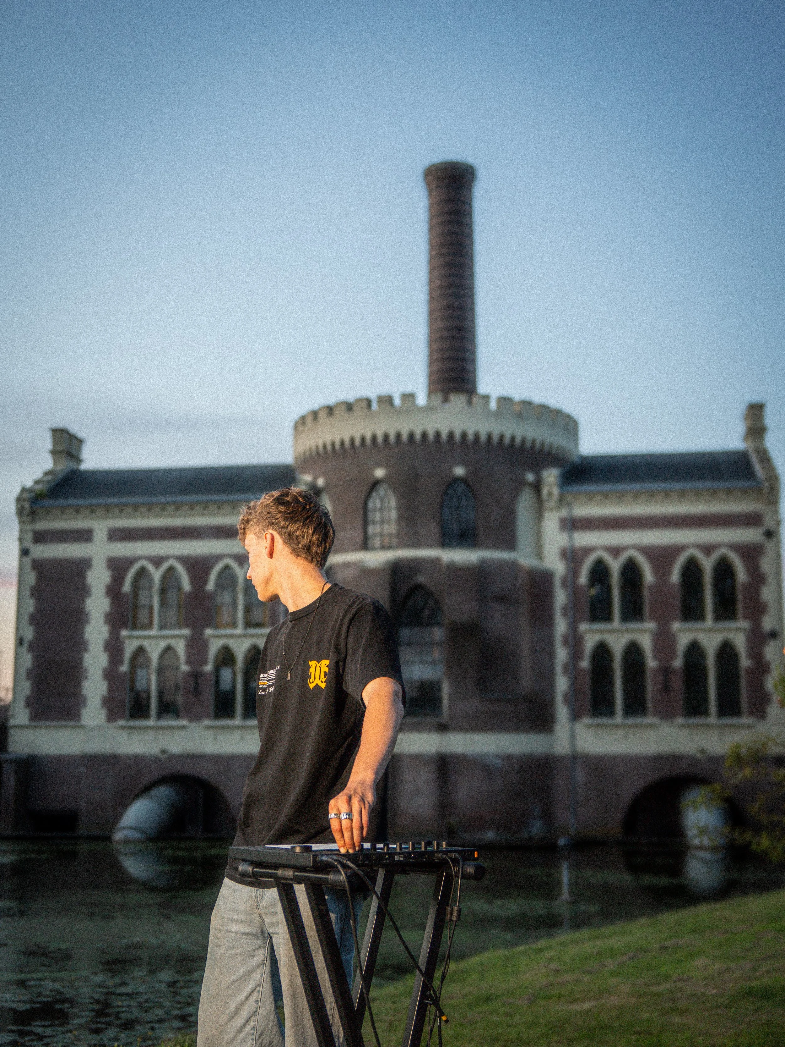 A person standing outdoors by a large historic building with arched windows and a tall chimney, holding a music keyboard on a stand.