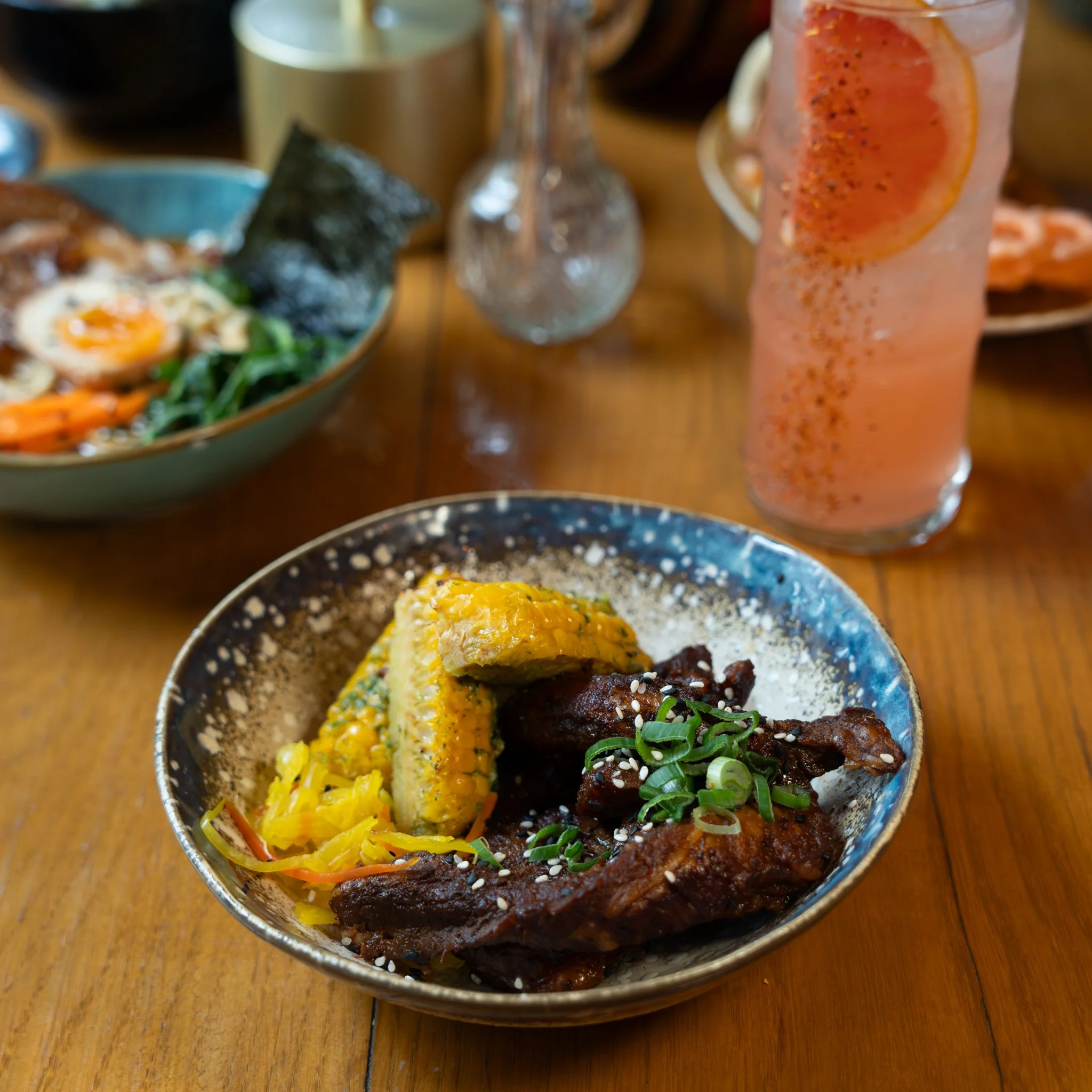 Asian-inspired dish with corn, greens, and meat in a bowl, alongside a grapefruit cocktail, on a wooden table.