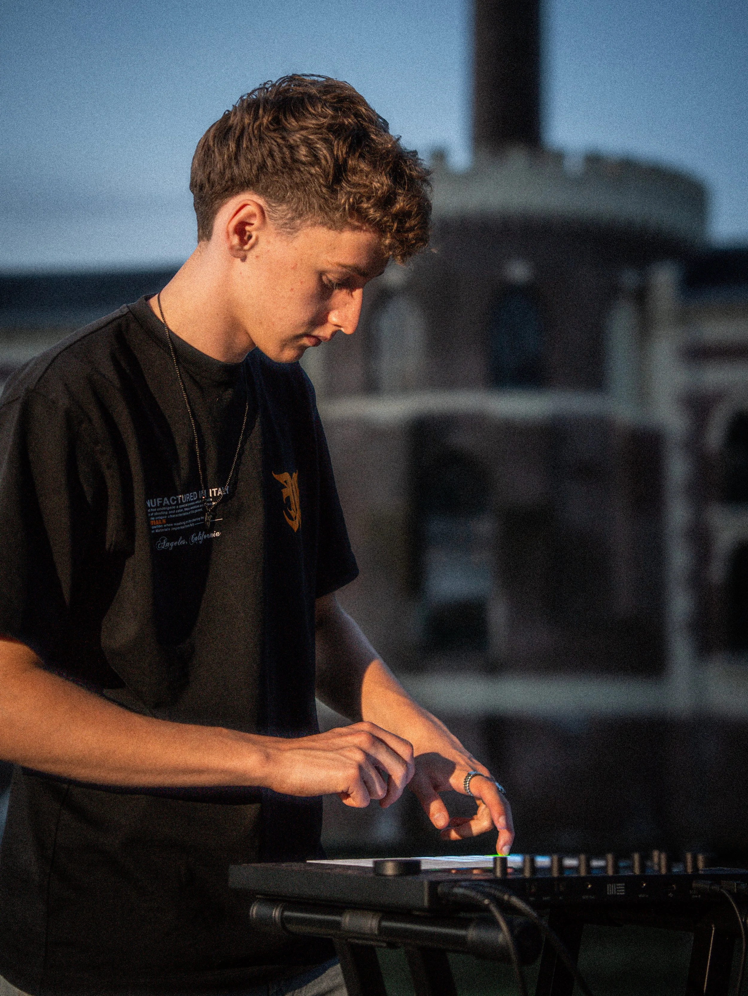 Young man playing a DJ mixer outdoors with a blurred building in the background.