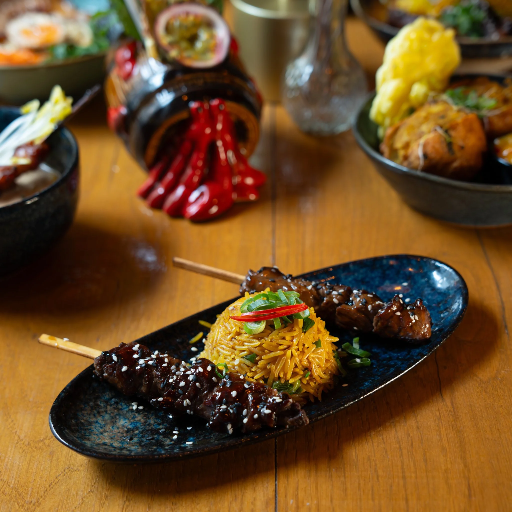 Plate of grilled meat skewers and seasoned rice on a wooden table, surrounded by other dishes and garnished with herbs and chili.