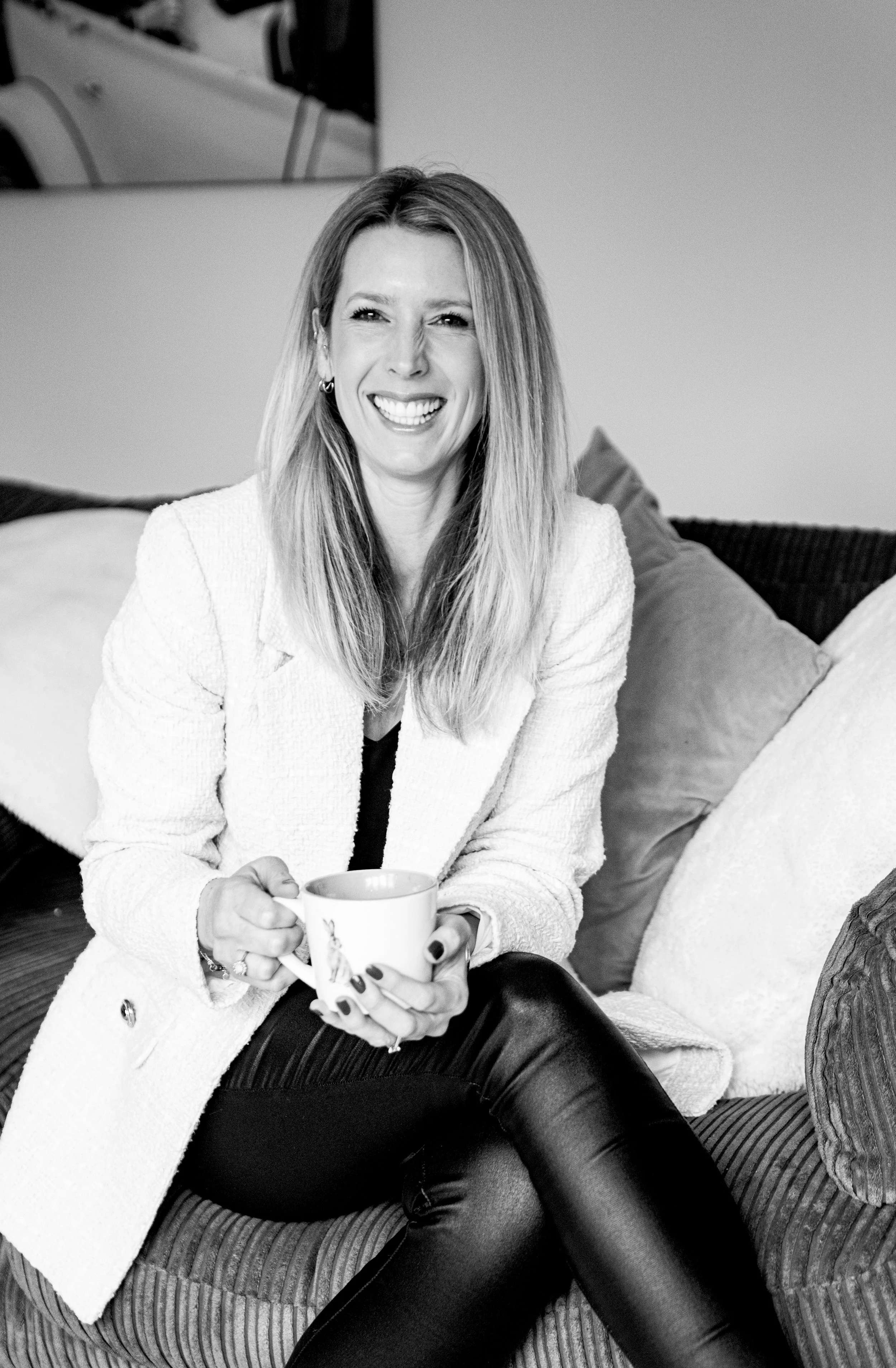A woman with long hair smiling and holding a mug, sitting on a couch with cushions in the background.
