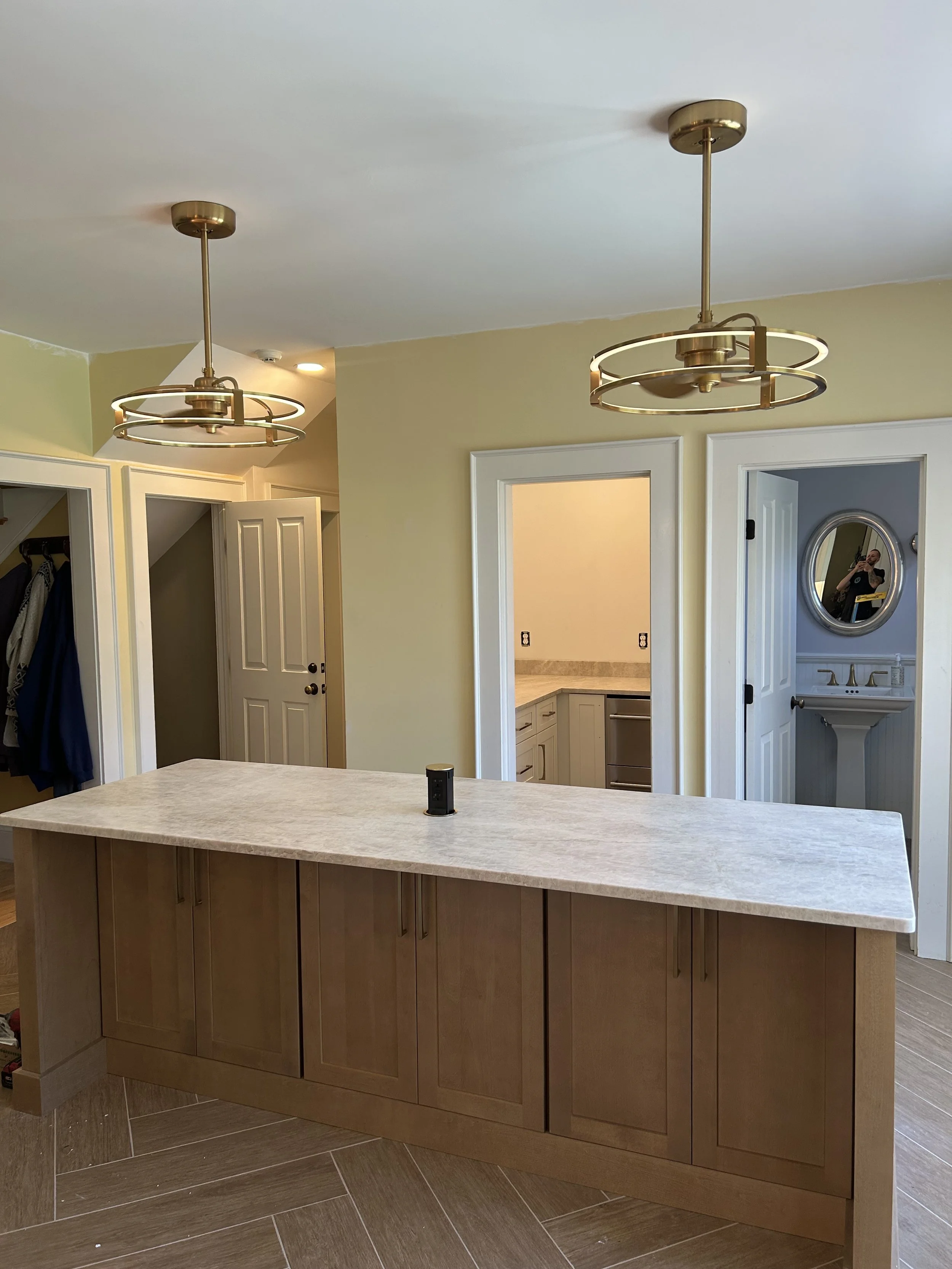 A kitchen island with a light-colored wooden base and a white countertop in a modern kitchen. Two gold and circular pendant lights hang from the ceiling. There are double doors and a small bathroom with a pedestal sink and oval mirror visible in the background.