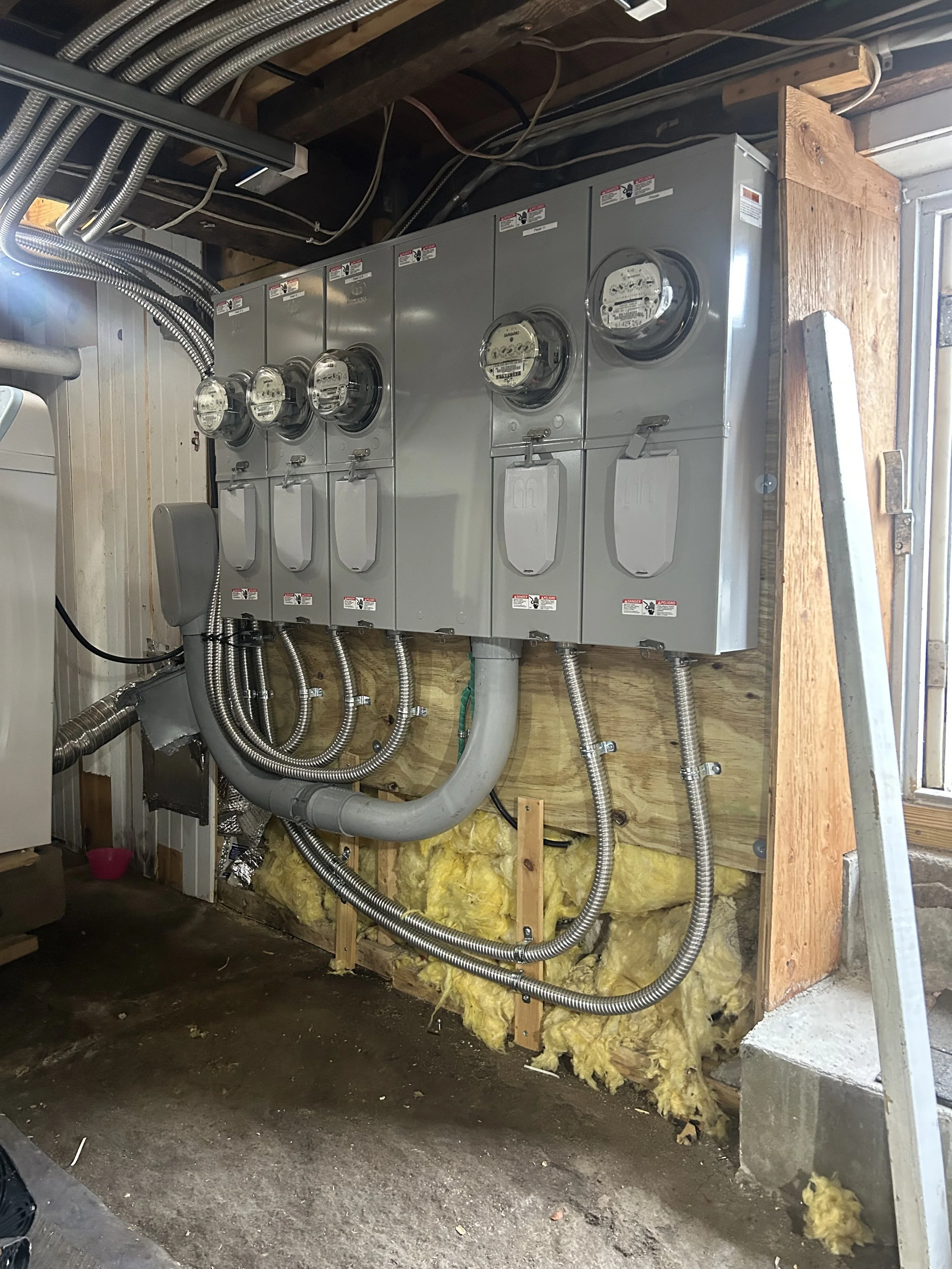 Electrical meter panel in a room under construction, with exposed insulation and wiring, and a ladder leaning against the wall.