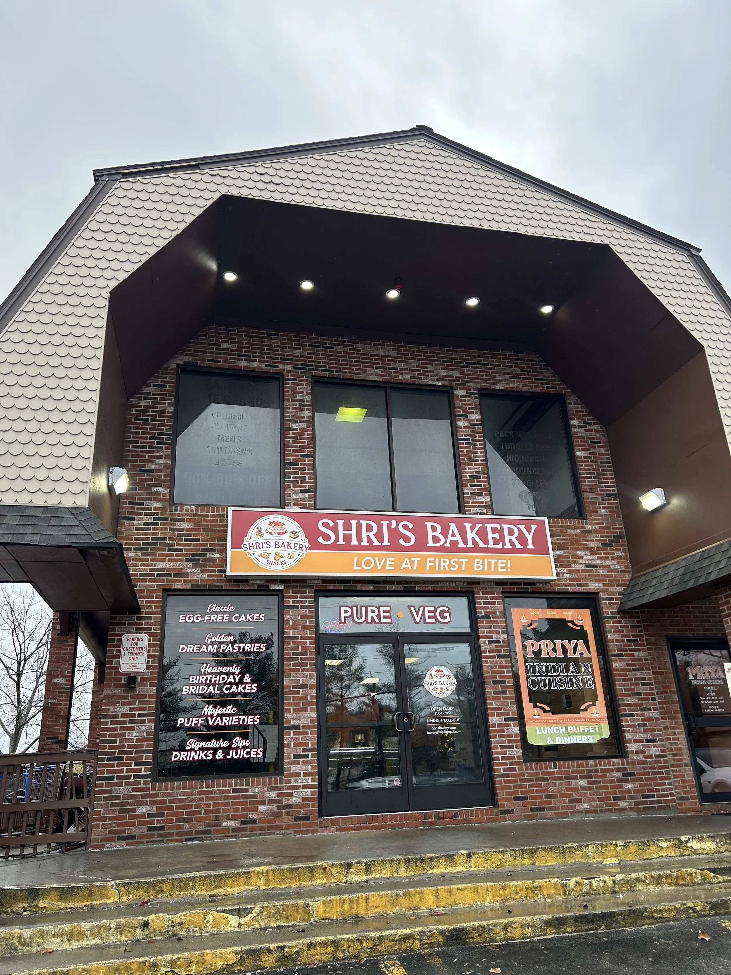 Exterior of Shri's Bakery, a brick building with black-framed windows and various signs advertising baked goods and Indian cuisine. There are steps leading up to the entrance, a cloudy sky, and some trees reflected in the windows.