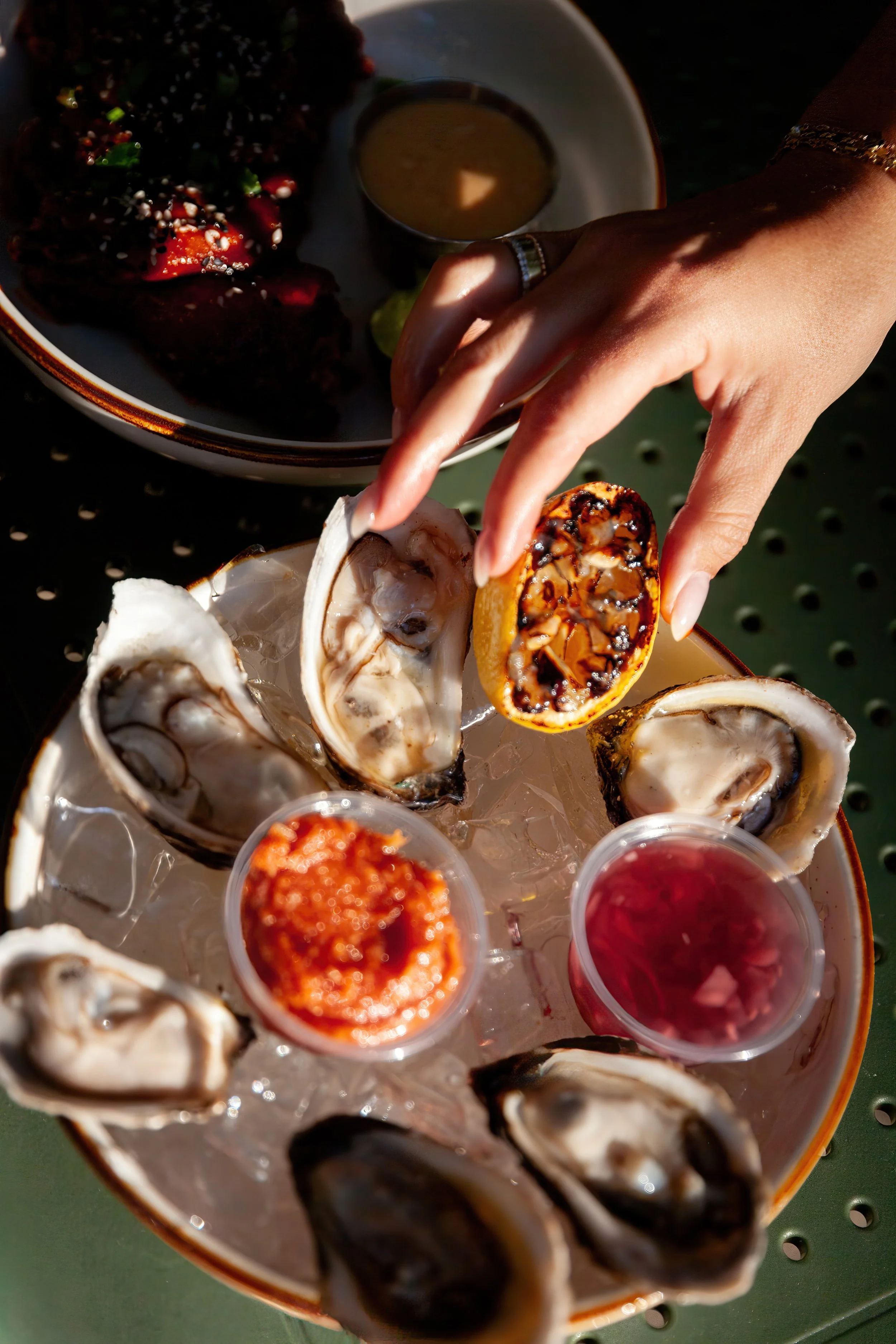 Close-up of a hand reaching for a grilled lemon half on a tray of oysters on ice, with some dipping sauces and a bowl of mustard in the background.