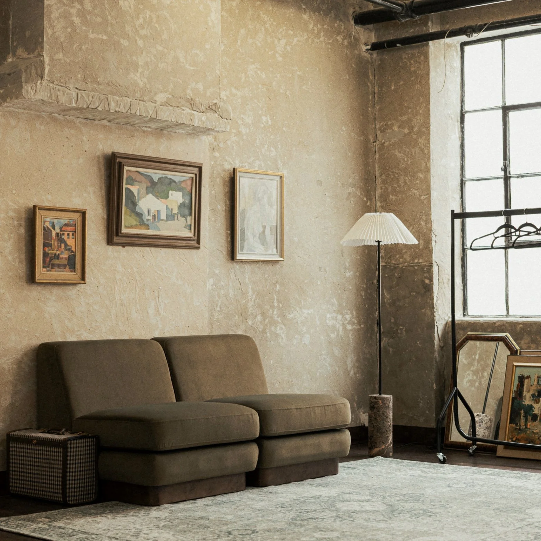 A vintage-style living room with a brown sofa, framed paintings on a textured beige wall, a tall floor lamp with a pleated shade, a large window with metal framing, a clothing rack with hangers, and framed artwork resting on the floor.