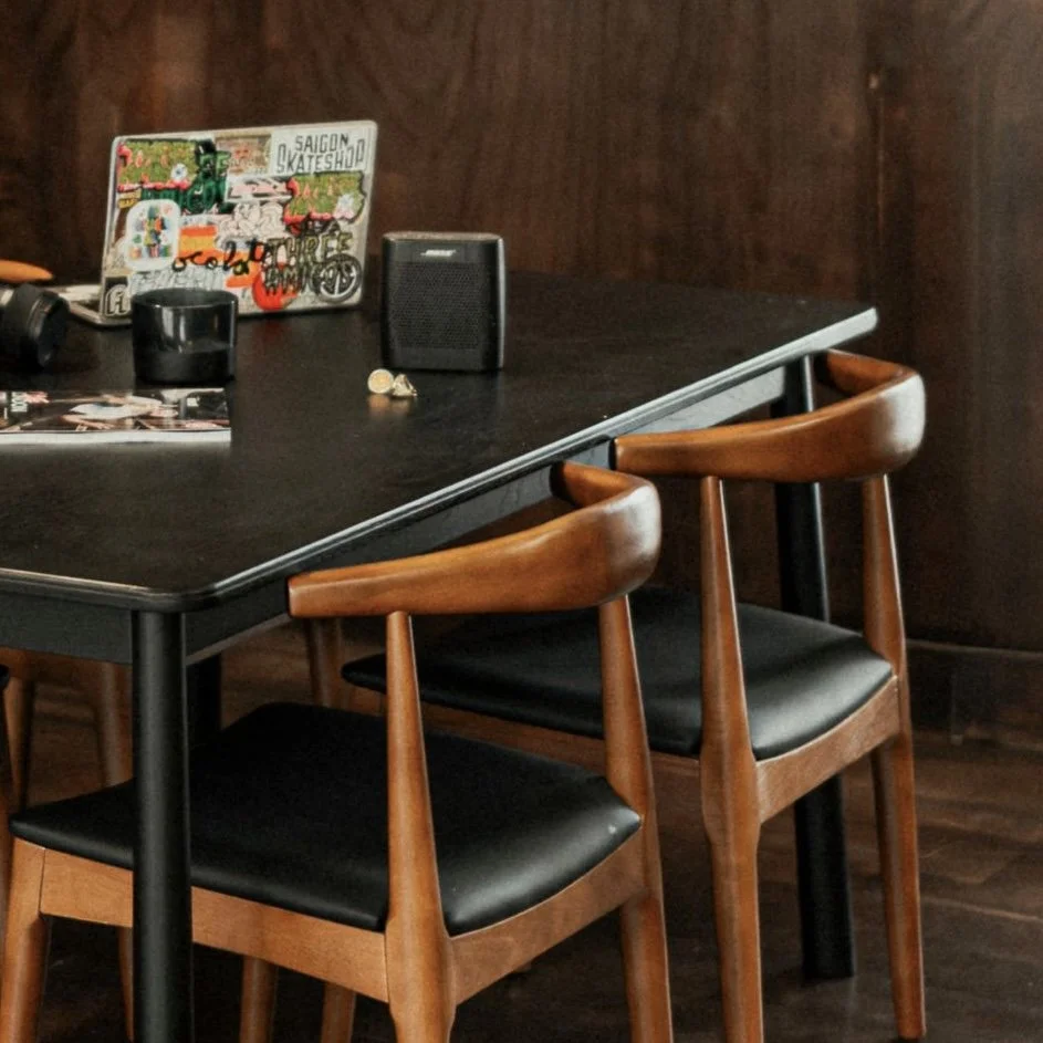 A black table with three wooden chairs, a laptop with stickers, a small speaker, a mug, earrings, and a magazine inside a room with dark wooden walls.