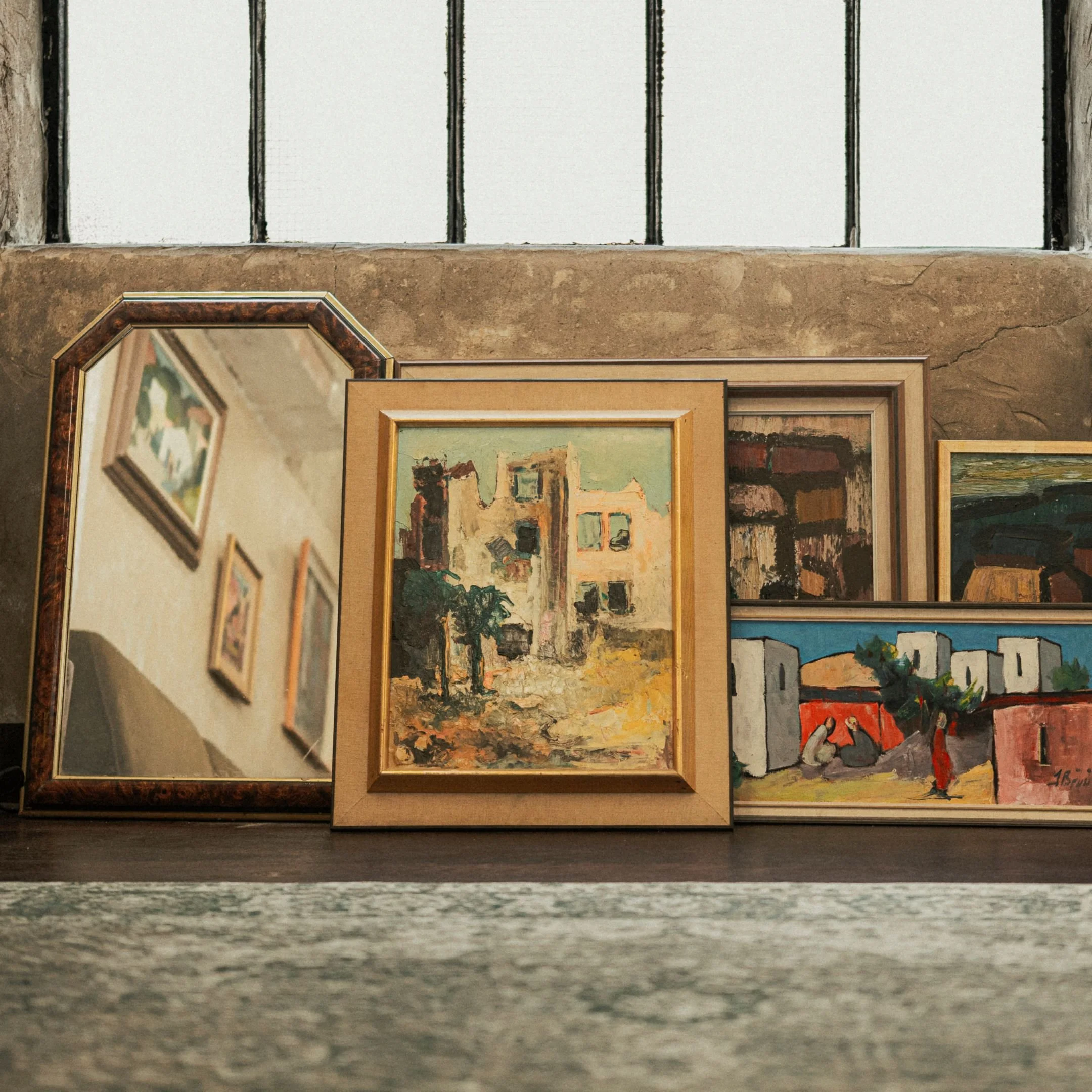 A collection of framed paintings and mirrors arranged on a surface against a brick wall, featuring various styles and landscapes.