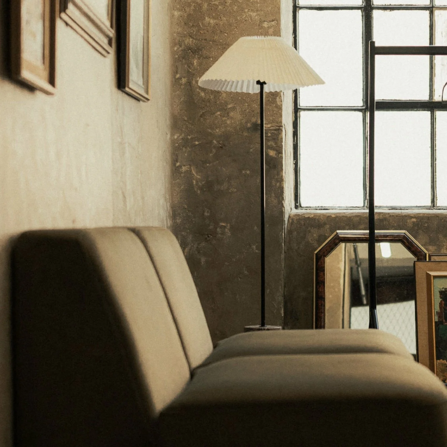 Cozy interior with a cushioned beige sofa, a tall vintage floor lamp with a pleated lampshade, an industrial-style window, and framed artwork on a textured wall.