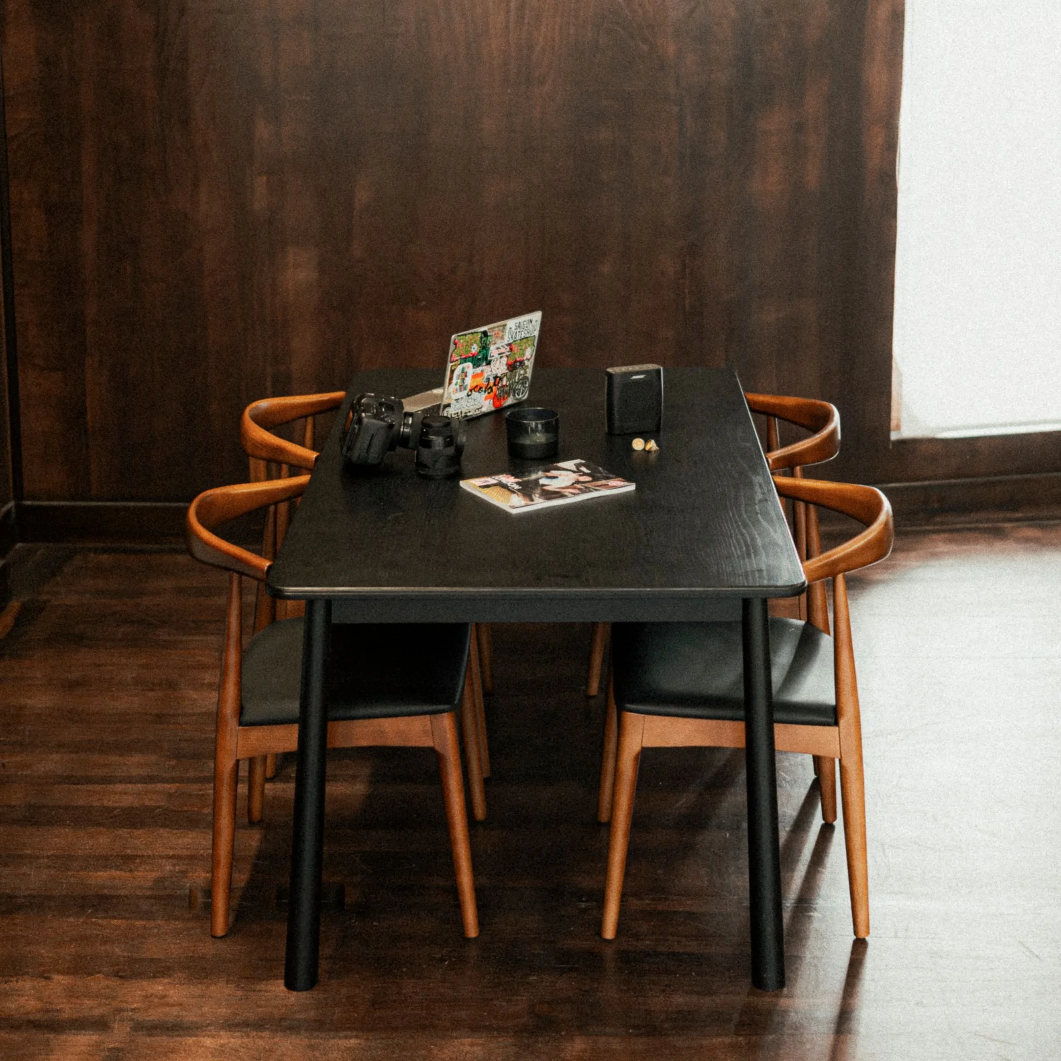 A black table with four wooden chairs around it, placed on a wooden floor and wall, with a laptop, camera, binoculars, a small speaker, and some magazines on top.