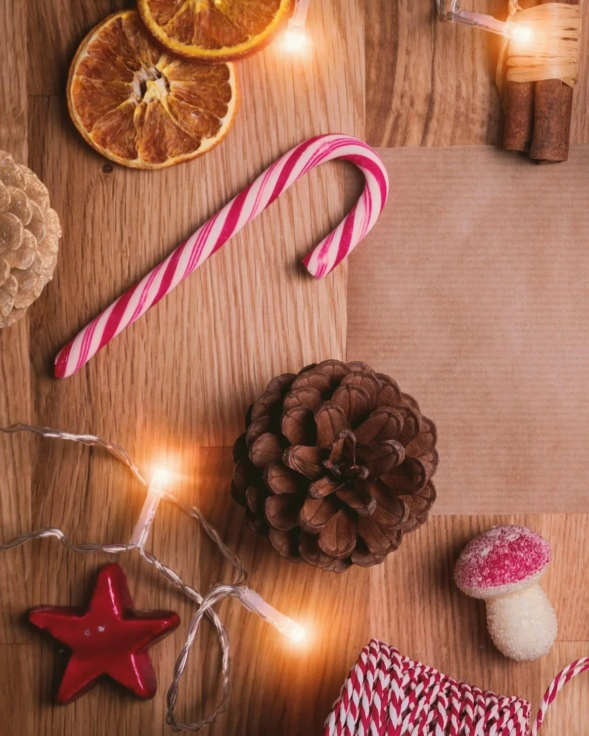 Decorative holiday scene on a wooden surface with dried orange slices, a red and white candy cane, a pine cone, string lights, a red star ornament, a sugar-coated mushroom-shaped candy, and red and white striped ribbon.