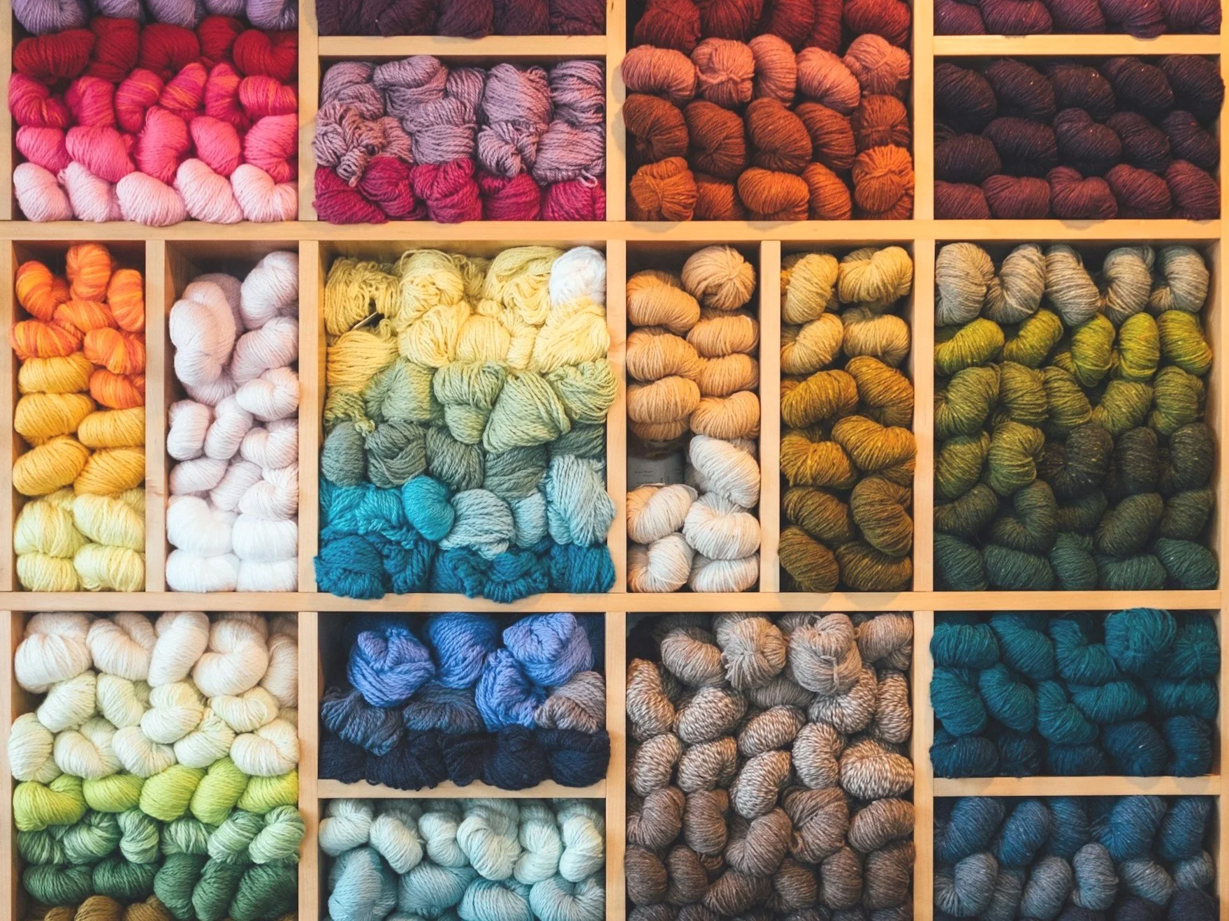 Colorful skeins of yarn organized in a wooden shelf, sorted by color in various shades, including reds, yellows, blues, greens, and neutrals.