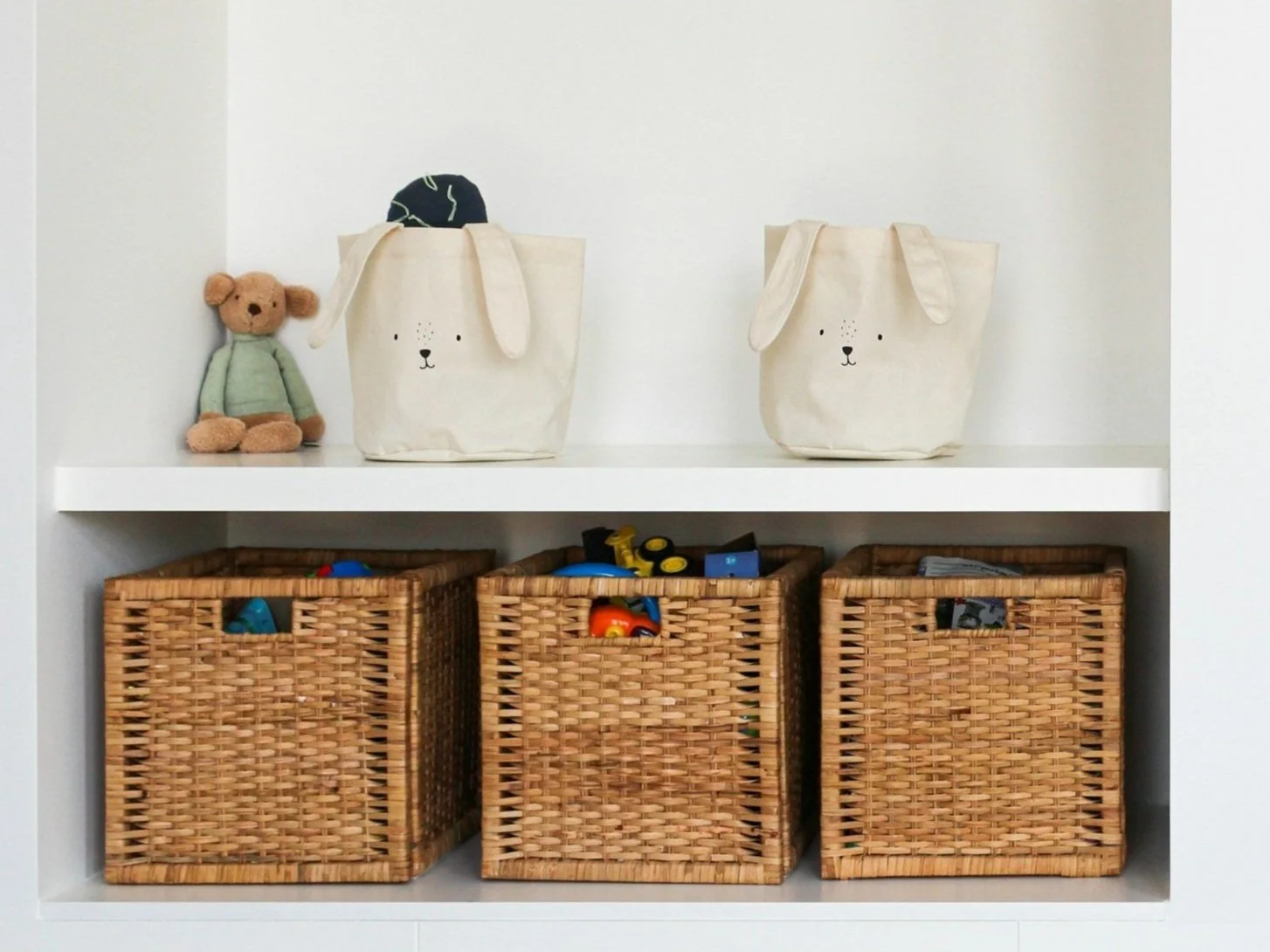Shelf with stuffed bear, fabric bags with rabbit faces, and three wicker baskets.
