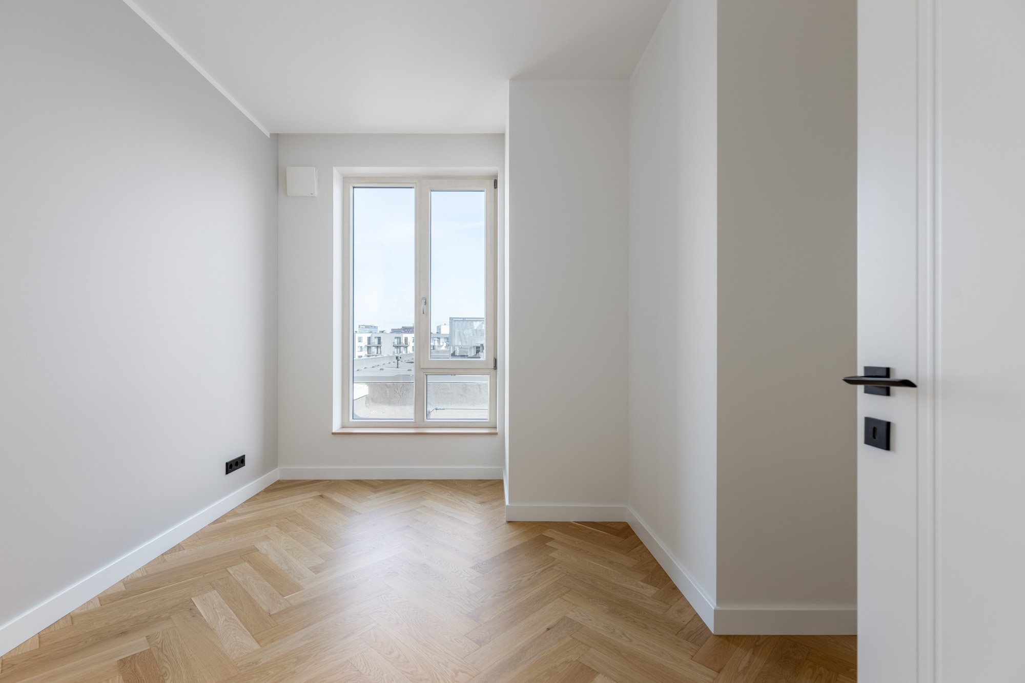 Empty room with a window showing a cityscape, wooden floor, white walls, and black electrical outlets.