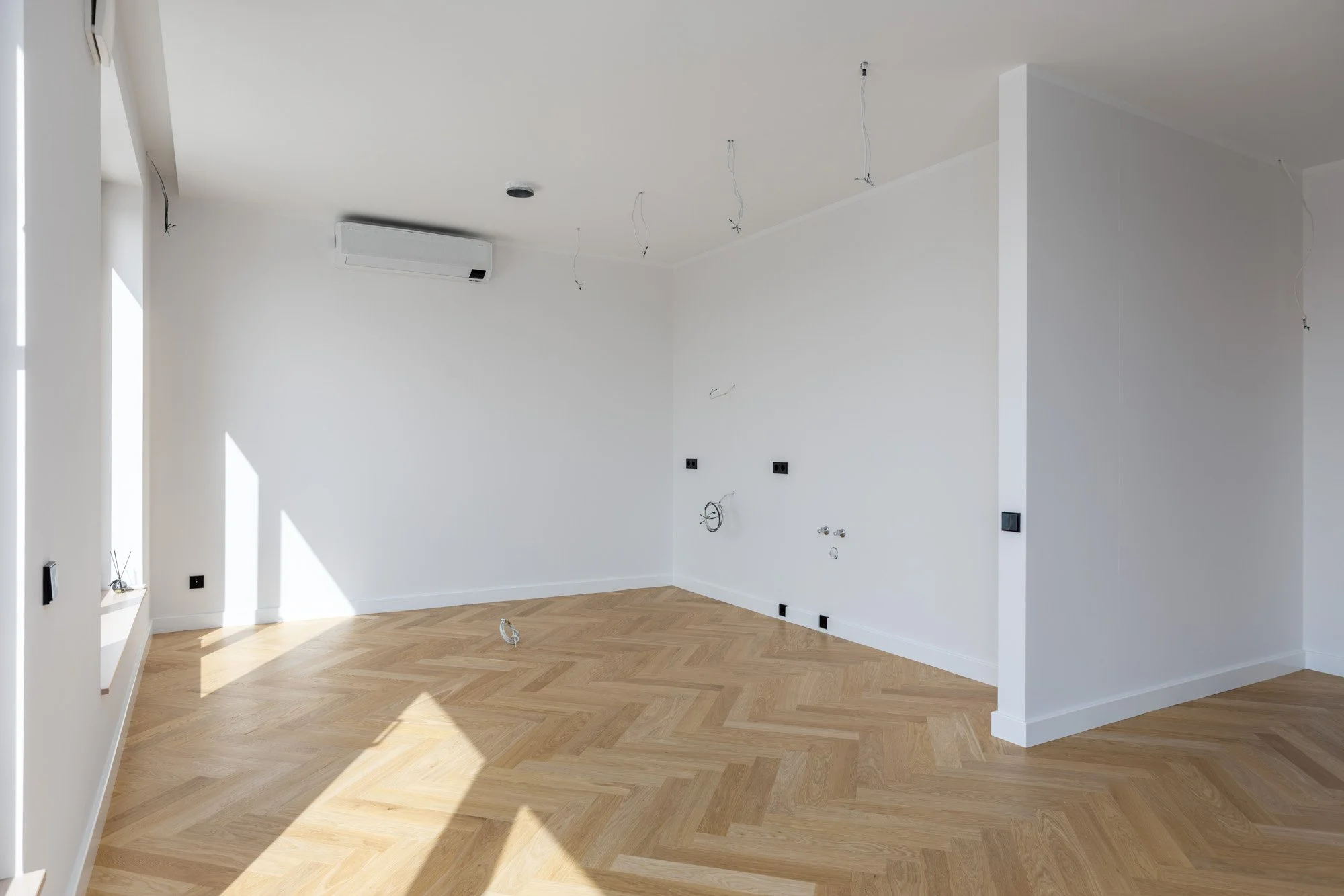 Empty room under construction with white walls, wooden flooring, and exposed electrical wiring on the ceiling.
