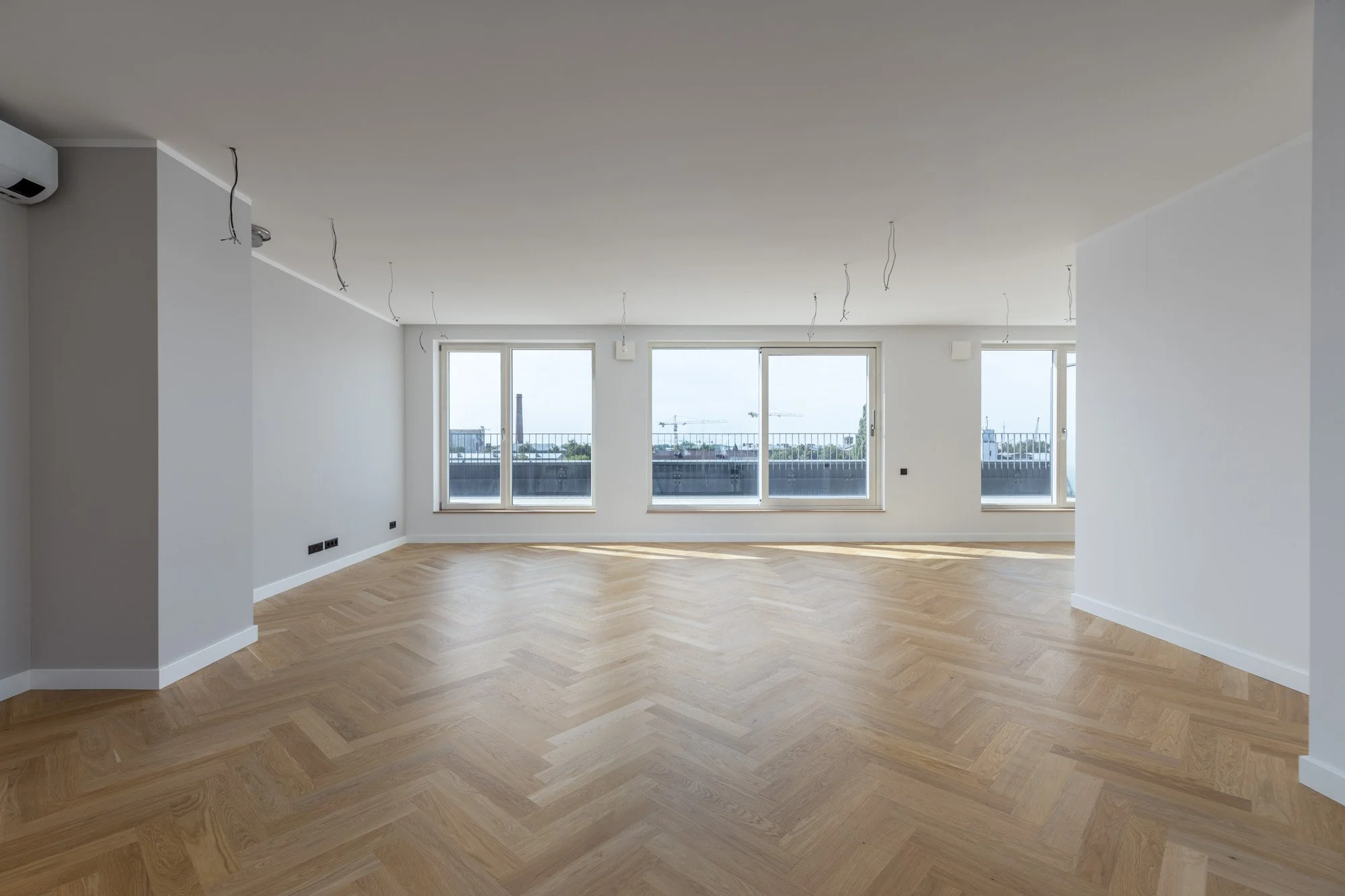 Empty room with light-colored wood flooring, white walls, and large windows showing a city view with cranes in the distance. Ceiling wires hang down, indicating ongoing construction or renovation.