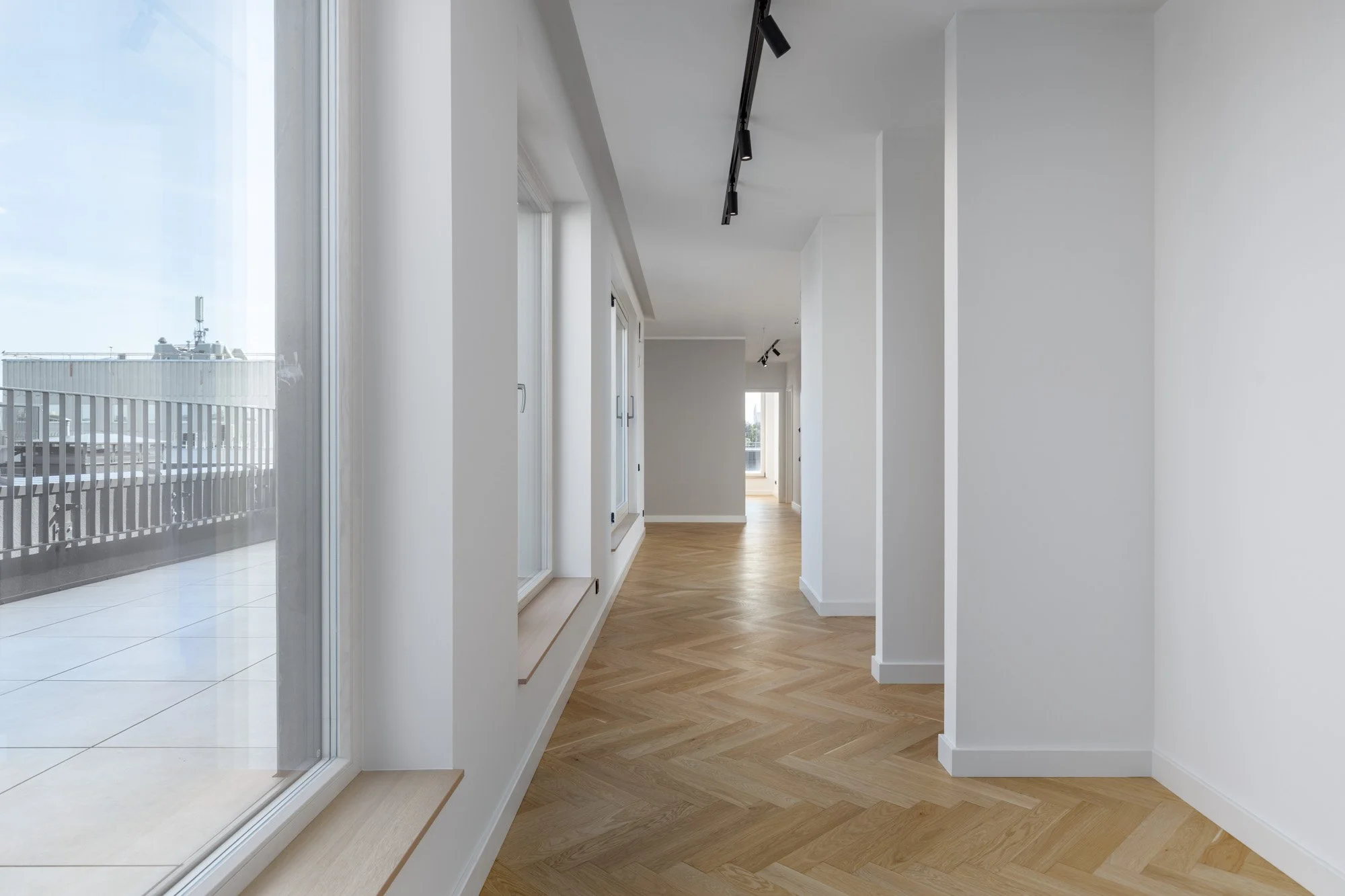 Empty modern hallway with large windows and balcony door, light wood herringbone flooring, white walls, and ceiling-mounted track lighting.