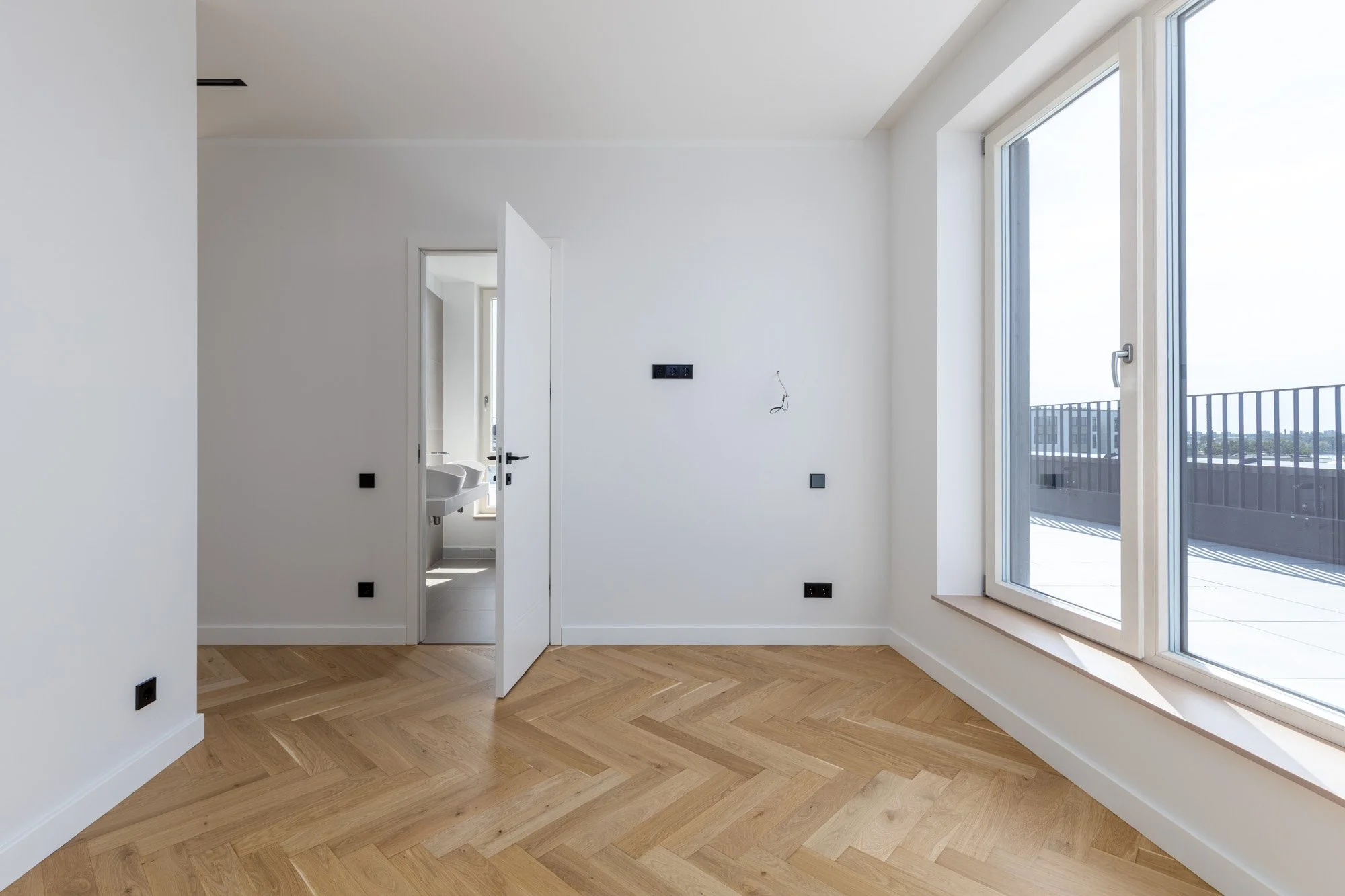 Empty modern room with large window, hardwood floor, and open door leading to what appears to be a bathroom.
