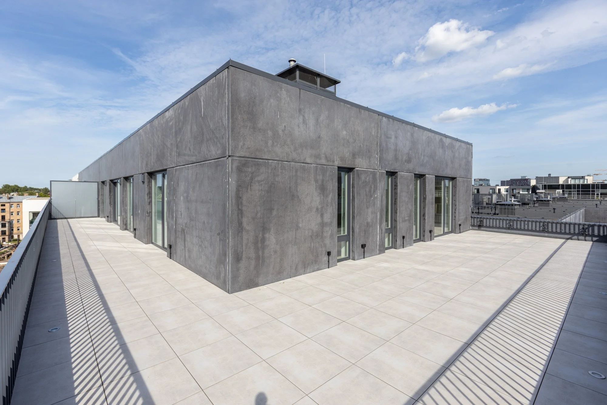 Modern gray building with large windows, rooftop terrace with tiled flooring, and city skyline in the background, under a partly cloudy sky.
