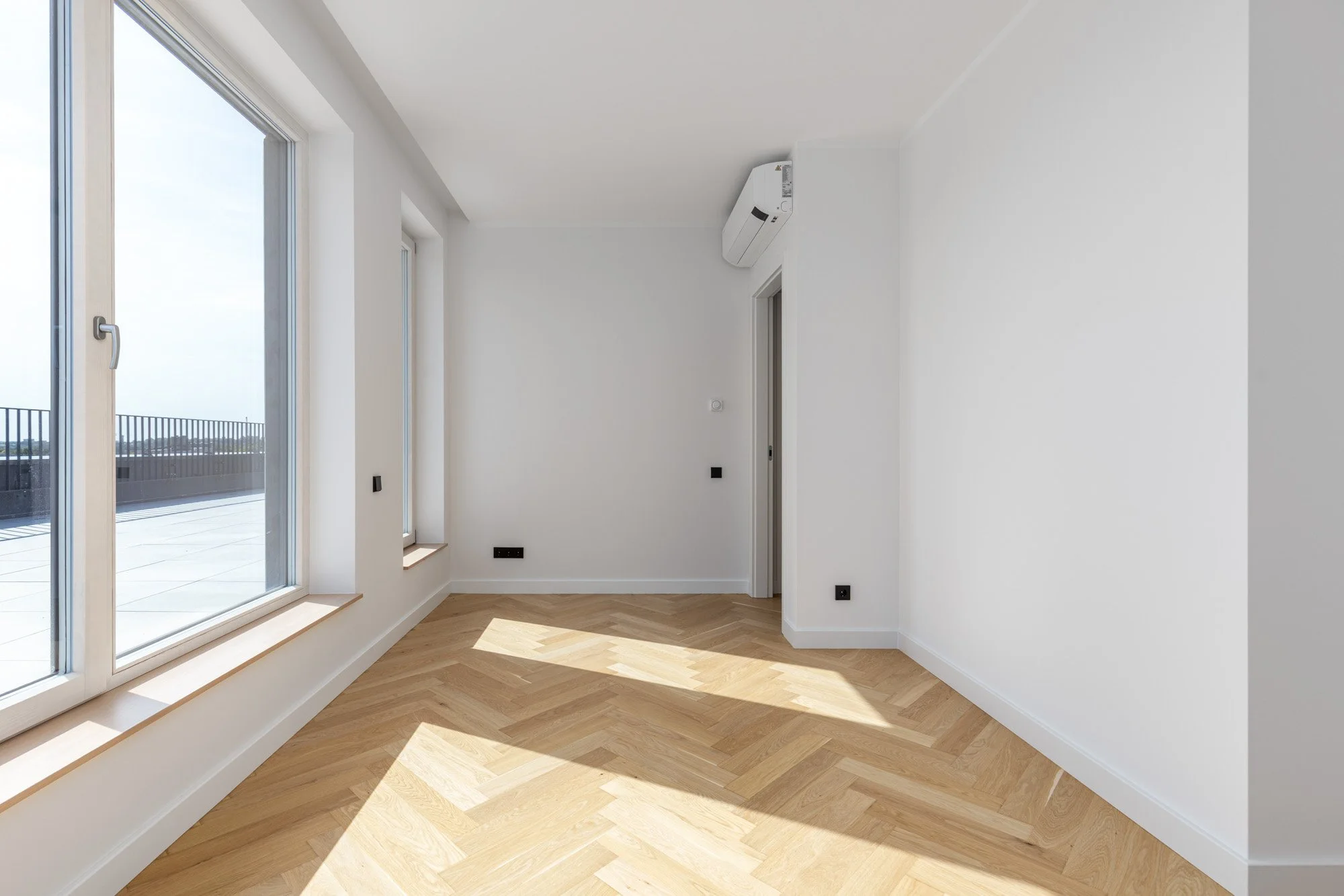 Empty room with white walls and large windows letting in natural light, hardwood floor, and a mounted air conditioning unit.