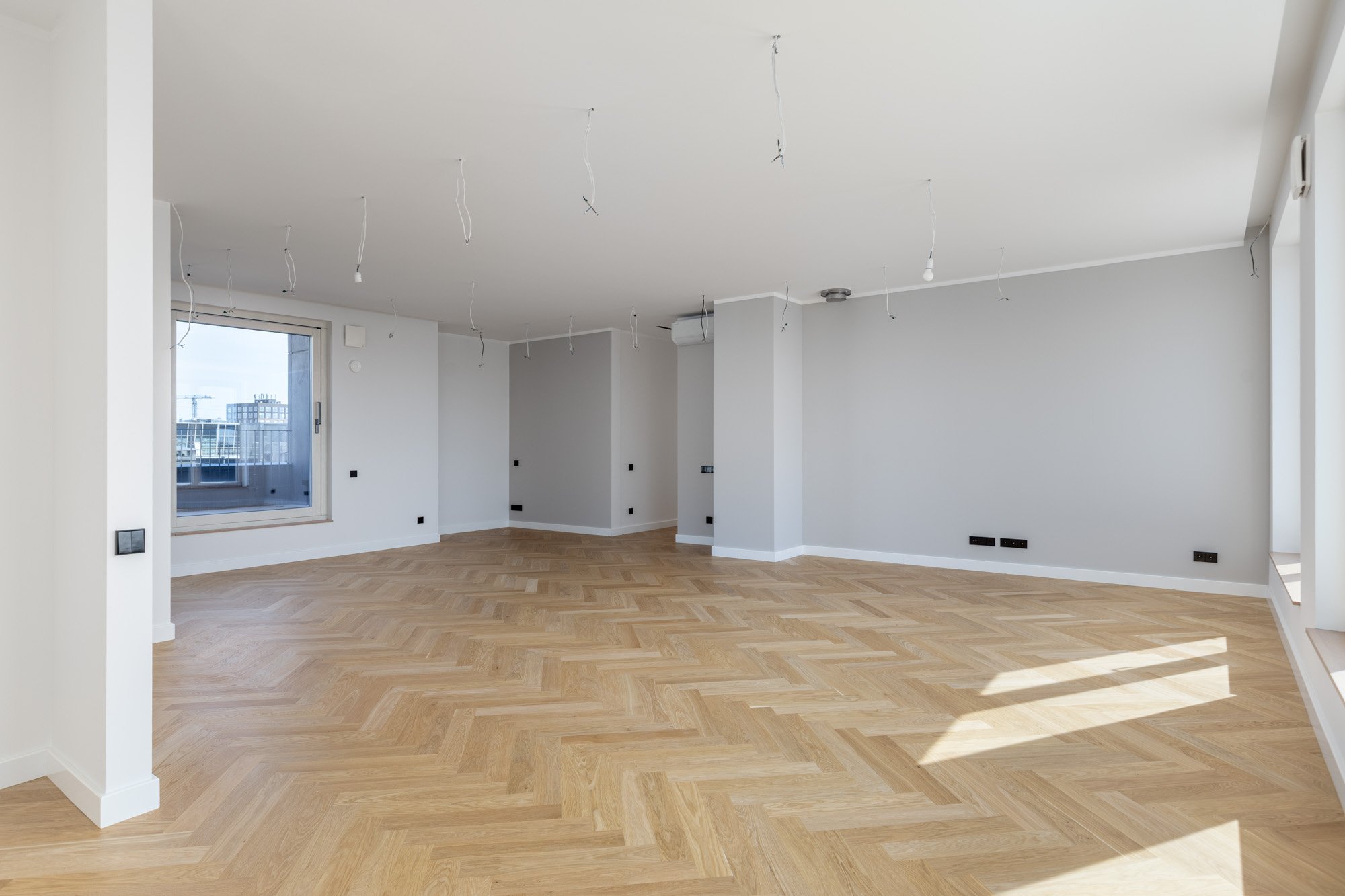 Empty room with white walls, light wood herringbone floor, large windows, and visible electrical and light fixtures hanging from the ceiling.